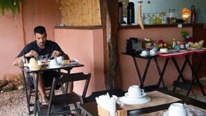 A man dining at an outdoor table with a basket of bread, a cup, and a plate of food. The table is set with white cups and saucers, and there is a small plant on the table. - Pousada Raízes (Alto Paraíso de Goiás)