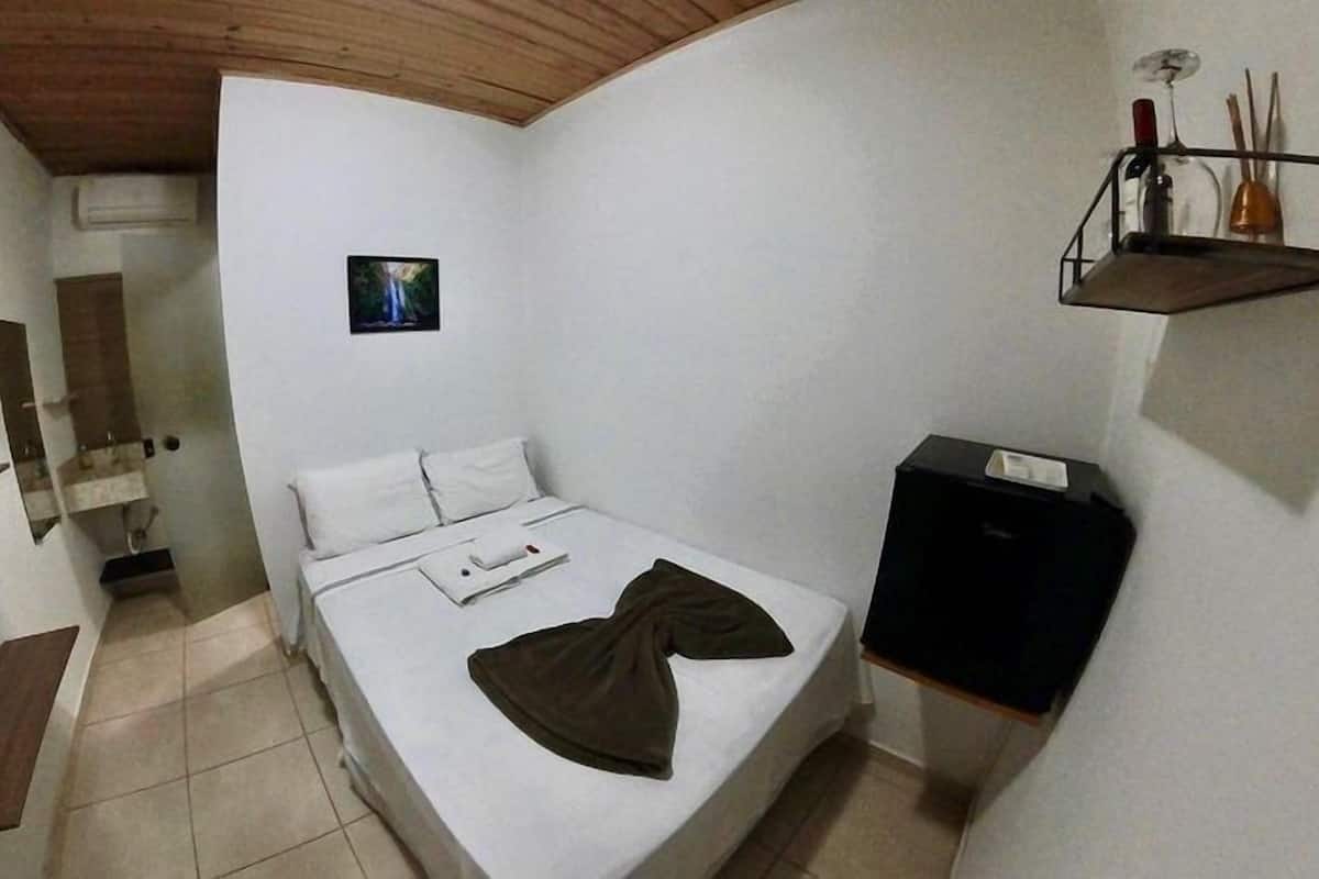 Une petite chambre avec un lit simple, un plafond en bois, une table de chevet, une petite étagère avec des objets décoratifs et un tableau encadré au mur.