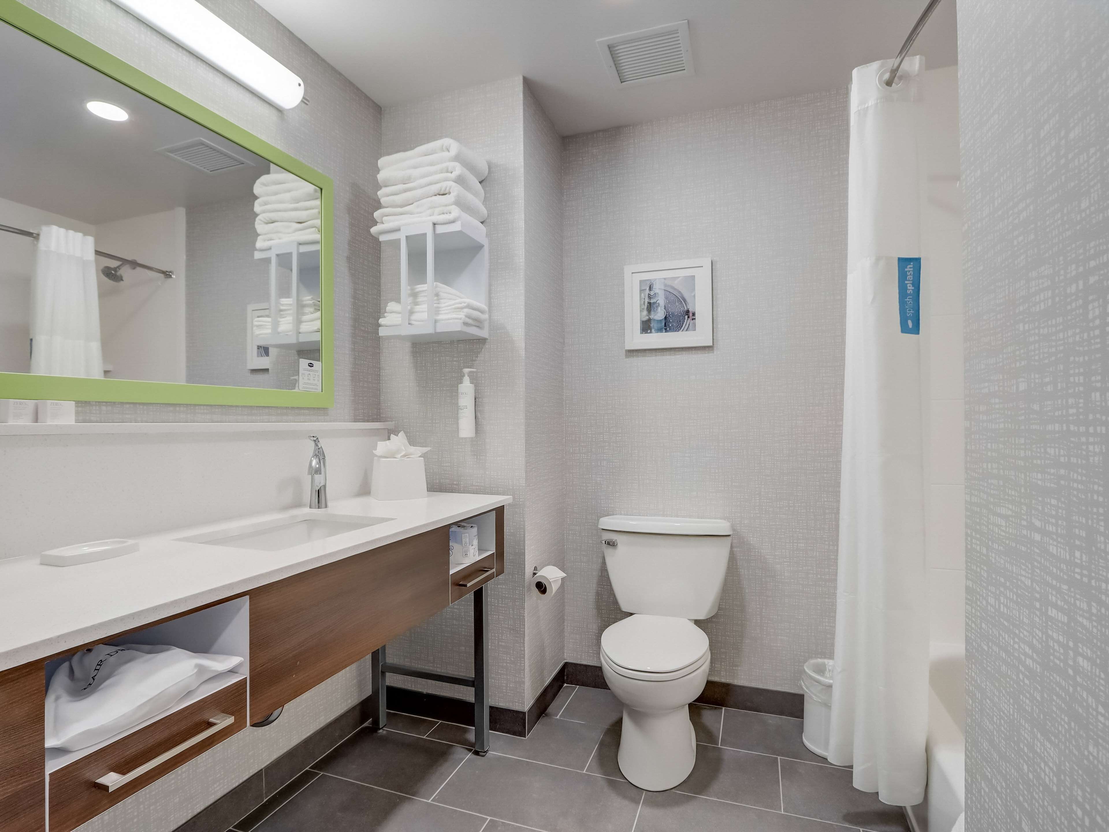 Une salle de bain moderne avec un lavabo blanc, un miroir et des toilettes. On y trouve des serviettes pliées sur une étagère, un tableau accroché au mur et un rideau de douche.
