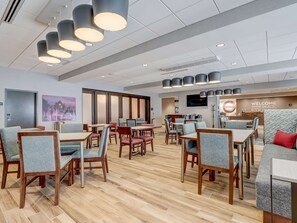 A modern hotel lobby with wooden flooring, multiple dining tables and chairs, and a reception area with a 'Welcome to the Eye Buck' sign.