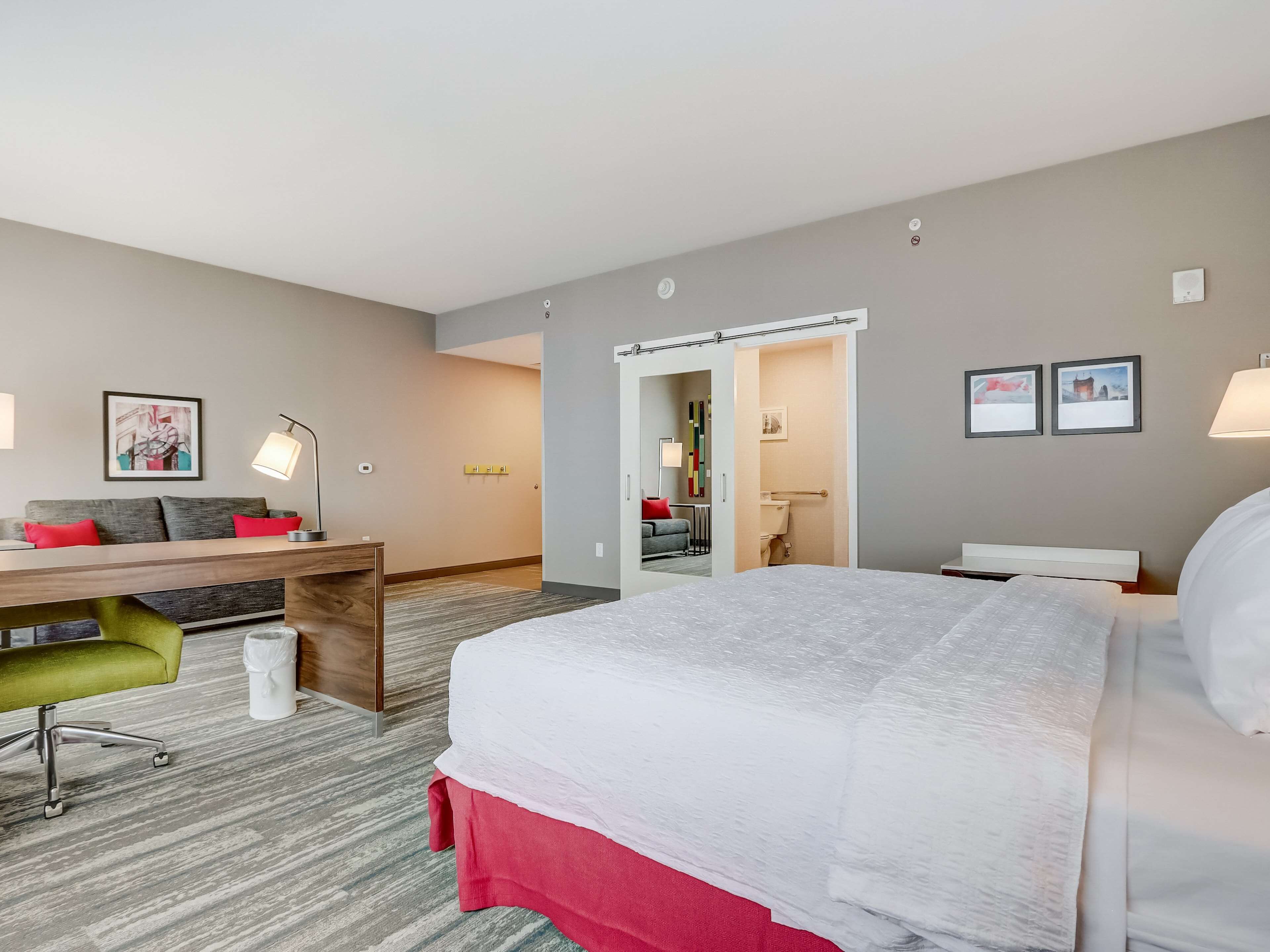 A hotel room with a large bed, a desk with a chair, a small table, and a sofa. There are two wall-mounted lamps, a picture on the wall, and a door leading to a bathroom.