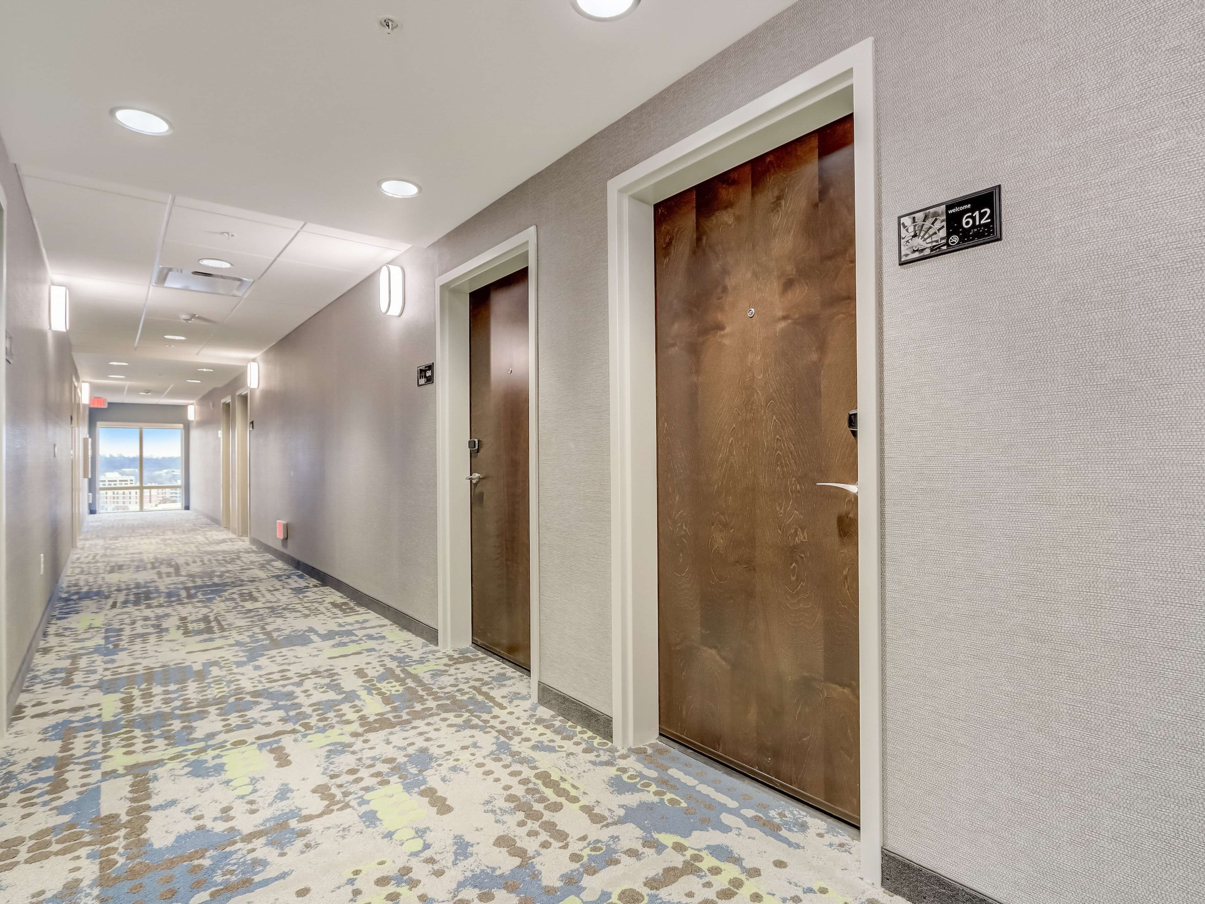 A hotel corridor with patterned carpet, wooden doors, and a view to the outside.