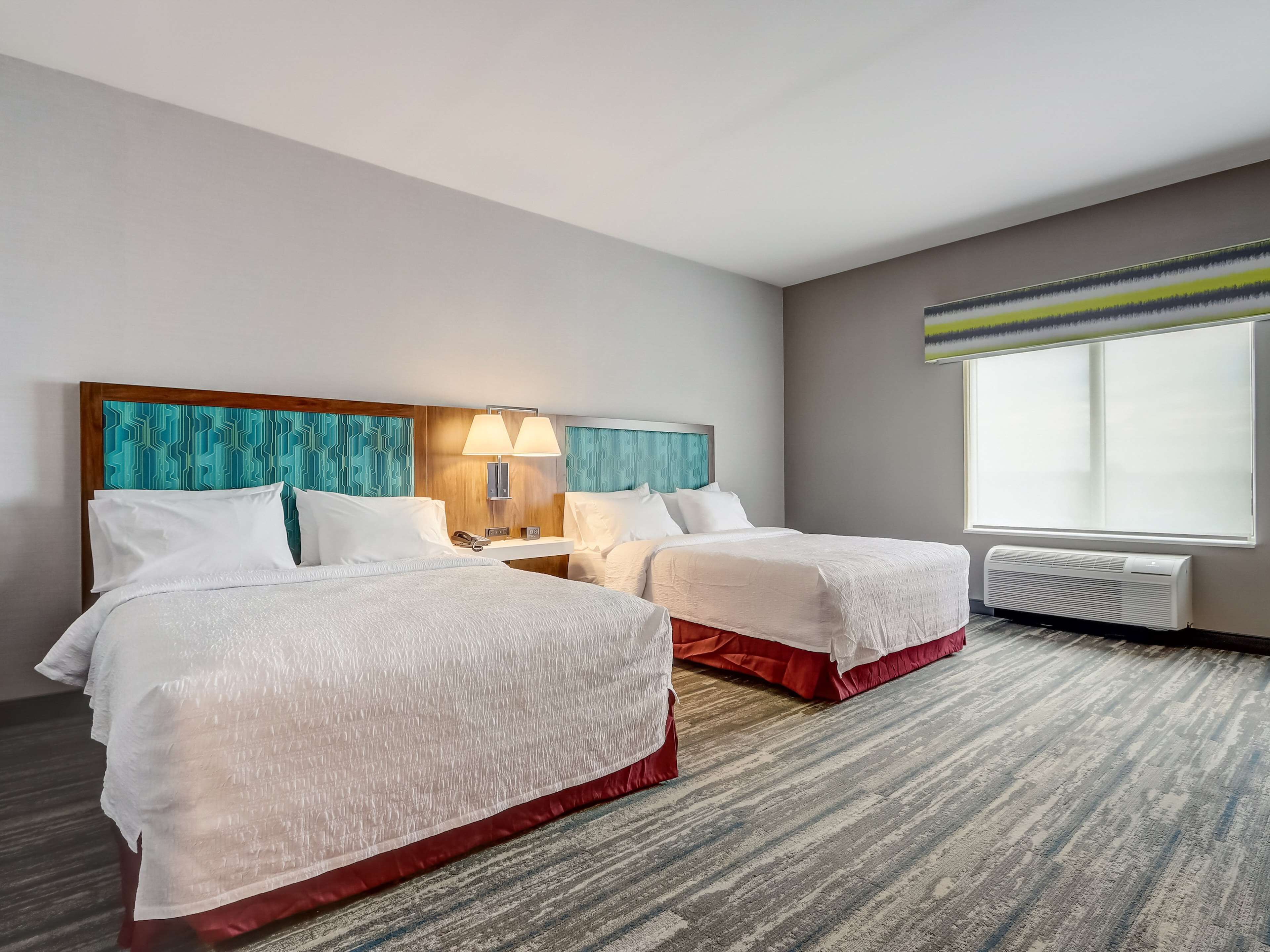 Une chambre d’hôtel avec deux lits, une grande fenêtre, un climatiseur fixé au mur et une tête de lit ornée d’un motif géométrique.