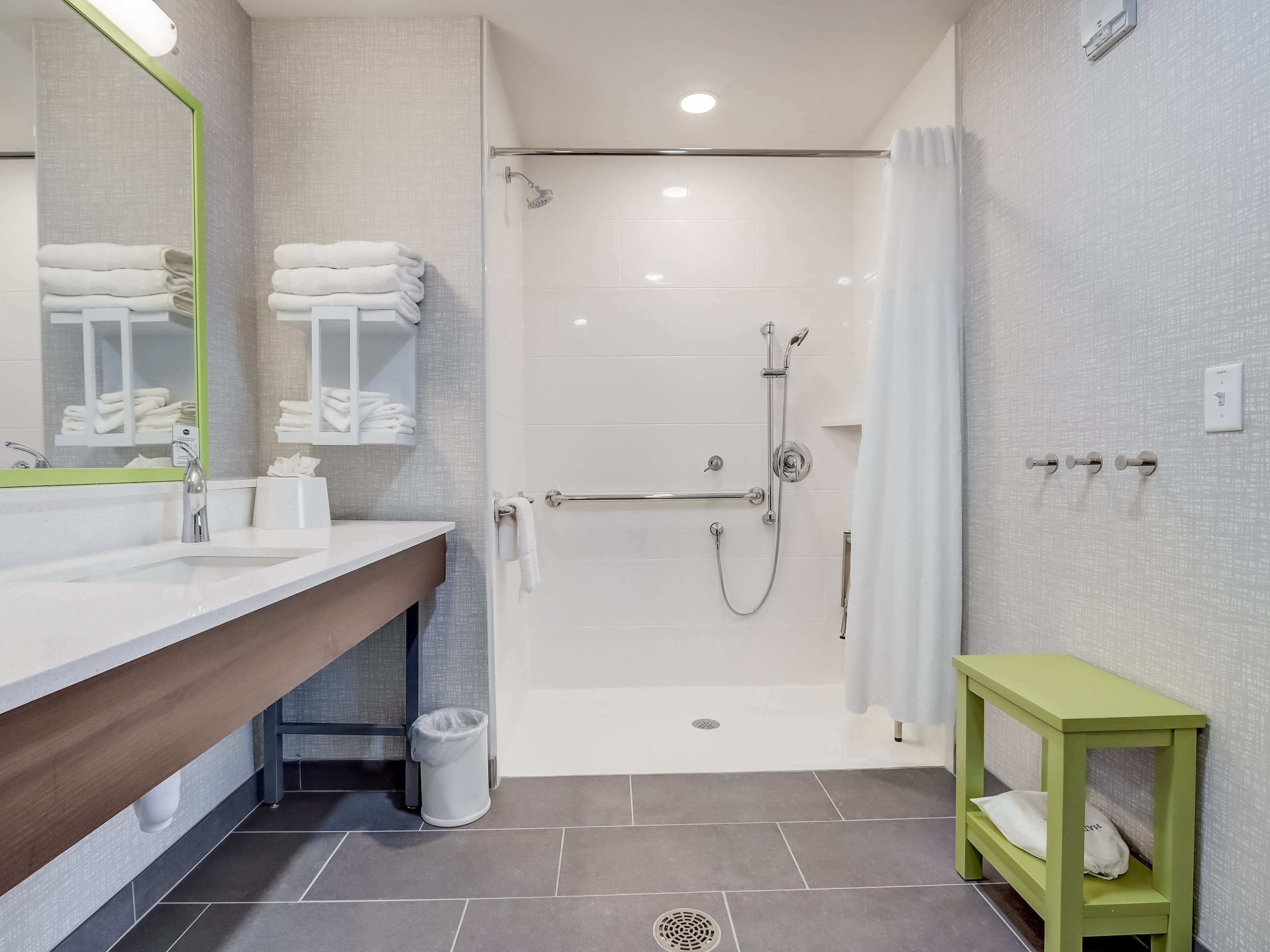 Un baño moderno con ducha, lavamanos y espejo, y un pequeño banco verde.