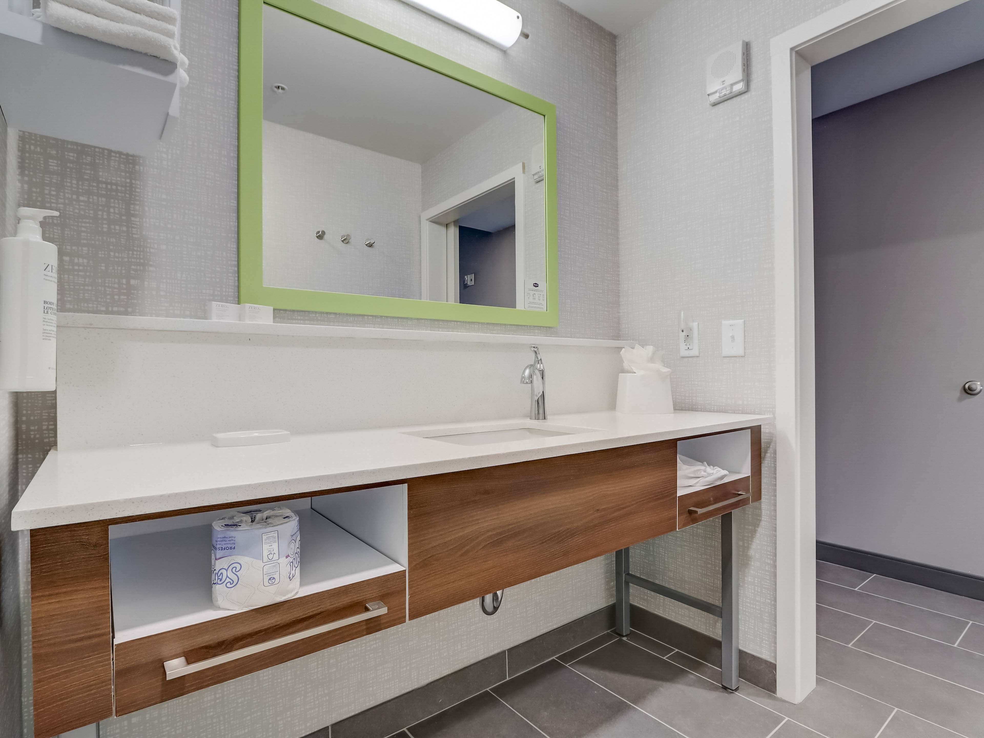 A modern bathroom with a white vanity, a green-framed mirror, a towel rack, and a toilet paper dispenser.