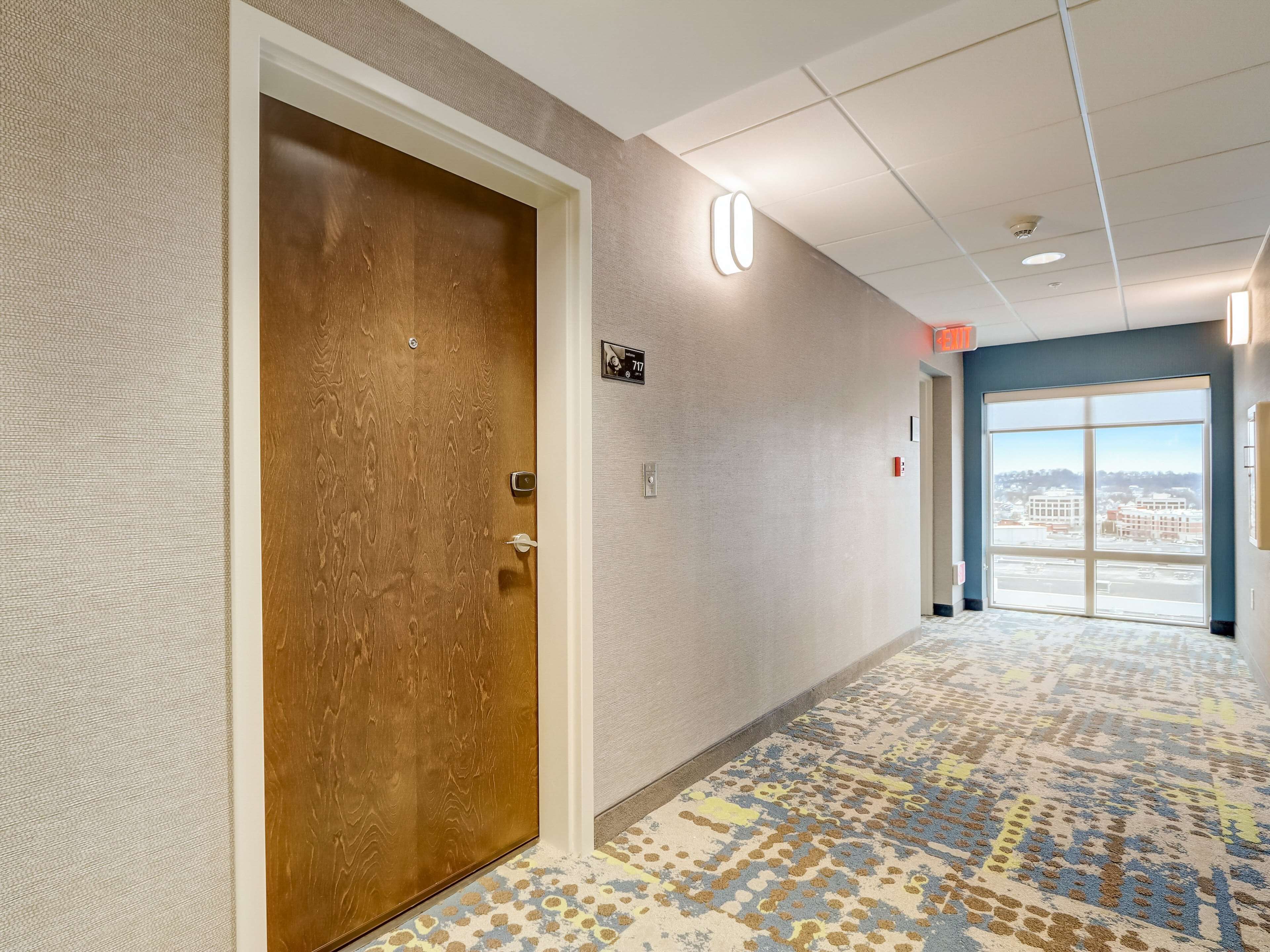 Un couloir avec une porte en bois, un mur à texture, une moquette à motifs et une grande fenêtre donnant sur la ville.