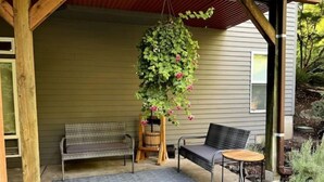 A covered porch with a bench and a chair, a hanging plant with pink flowers, and a small round table. - The Cellar @Lively Farm
a Family Friendly Hobby Farm Stay (Newberg)