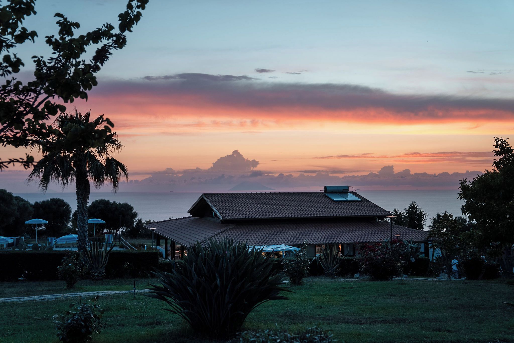 A house with a tiled roof, surrounded by a garden with palm trees and other vegetation, overlooking a body of water.