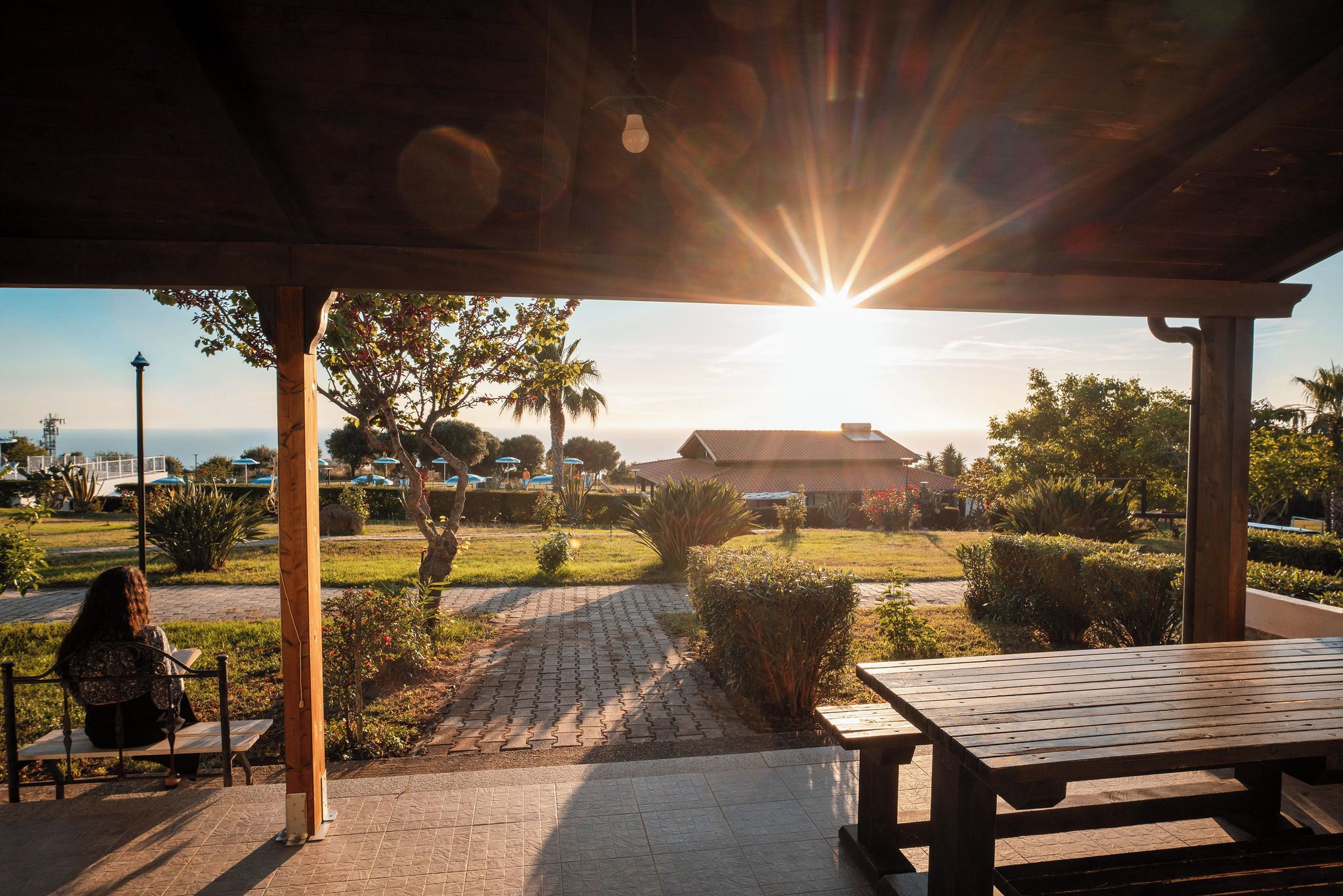 A covered outdoor area with a table and a bench, a paved pathway, and a view of a building and landscape.