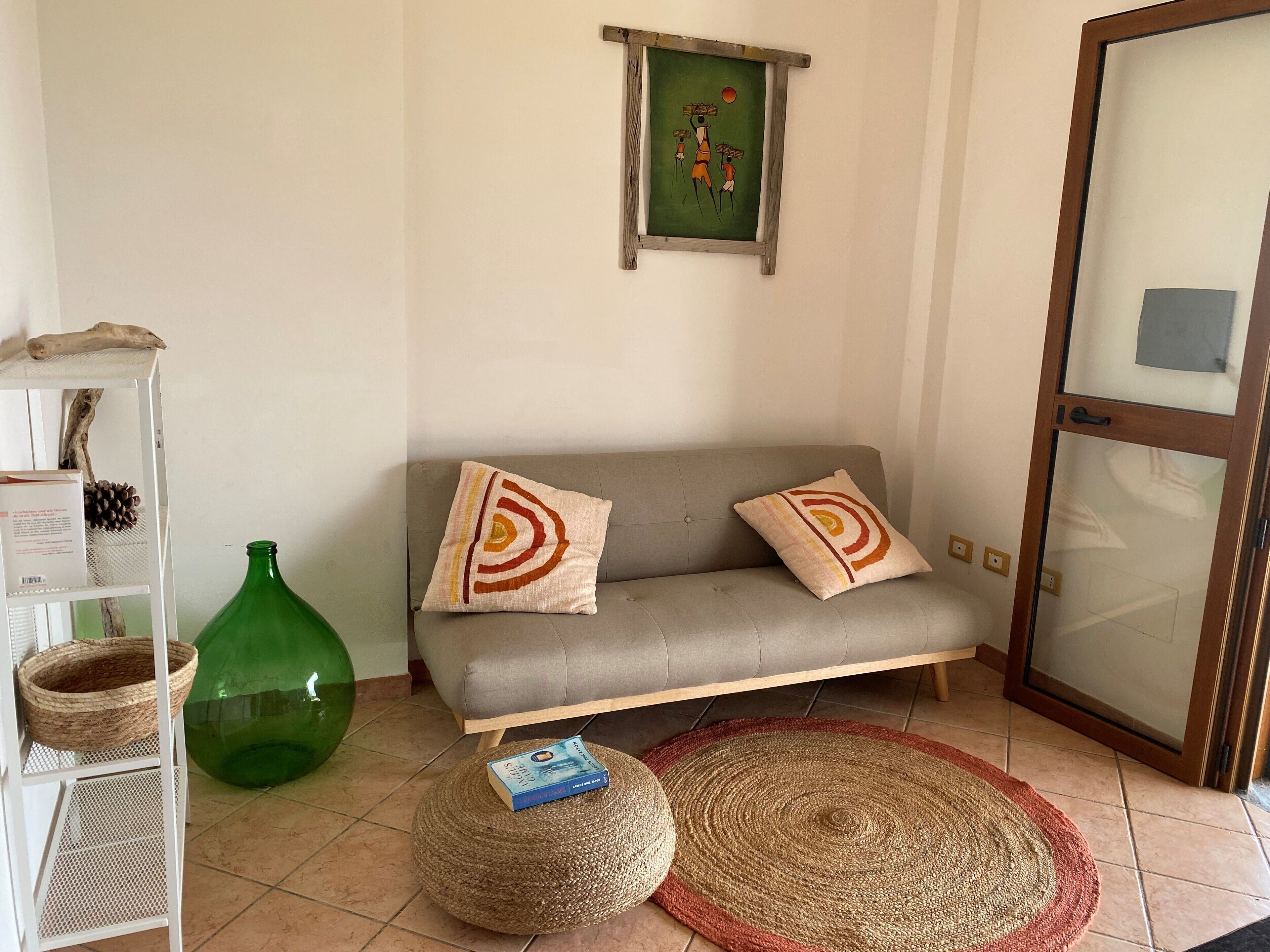 A living room with a grey sofa, two patterned cushions, a green vase, a woven pouf, a book, and a framed artwork on the wall.