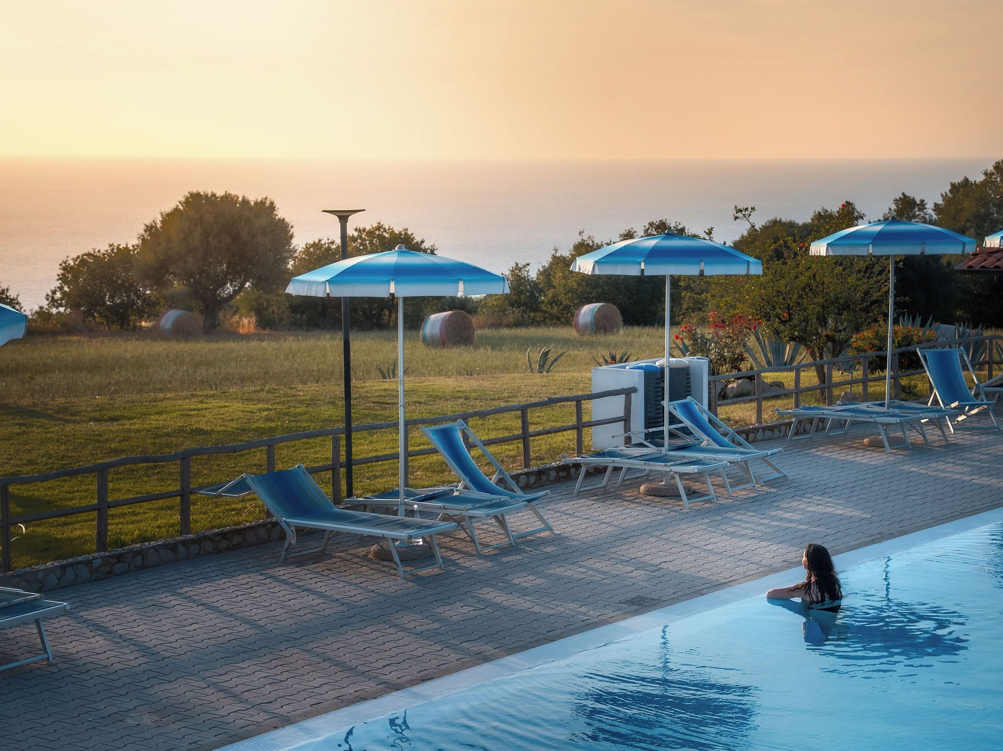 Un espace aménagé au bord de la piscine avec des chaises longues et des parasols, une personne dans la piscine et des bottes de foin en arrière-plan.