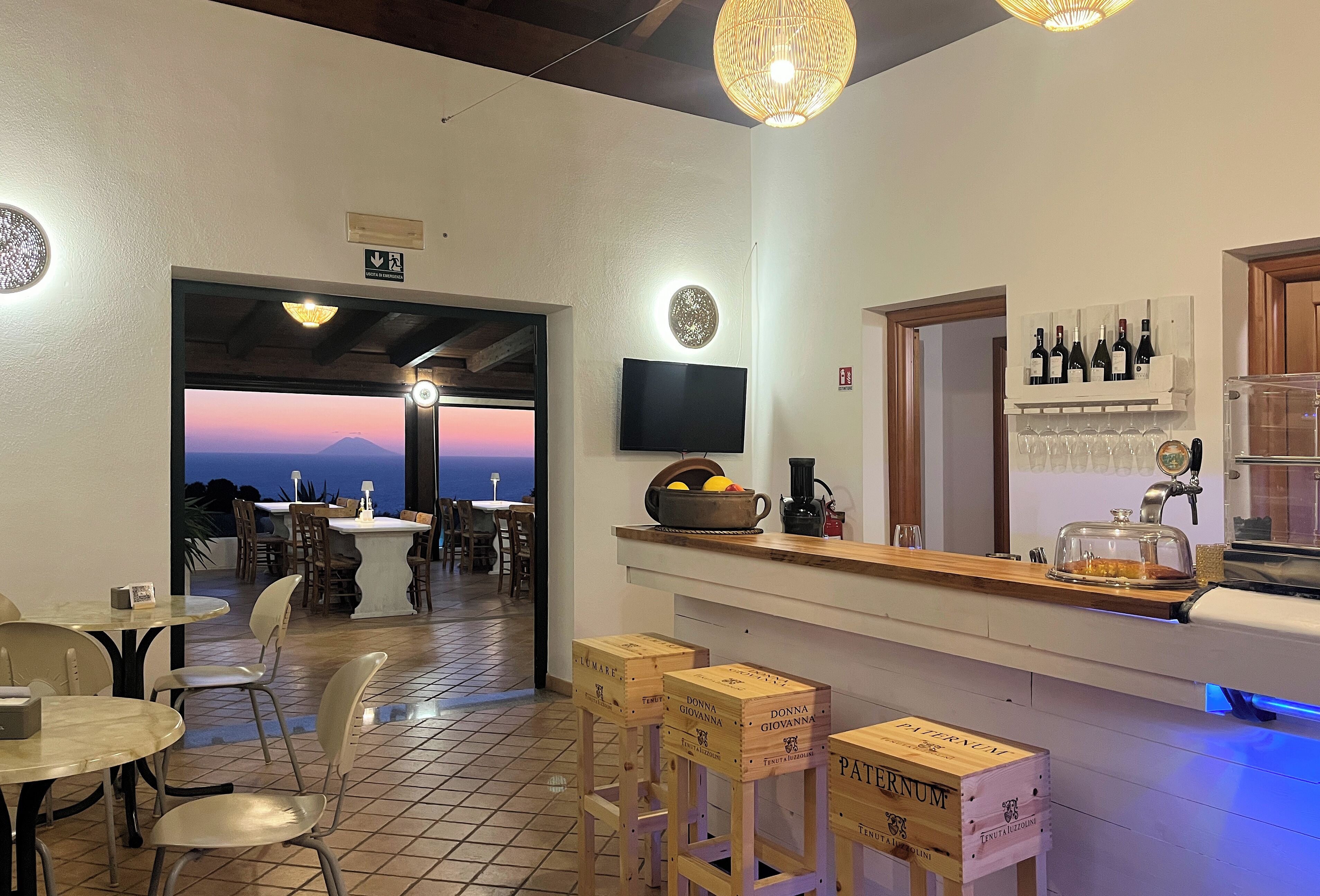 A restaurant interior with a bar area, wooden crates labeled 'PATERNUM', and a view of the sea through an open doorway.