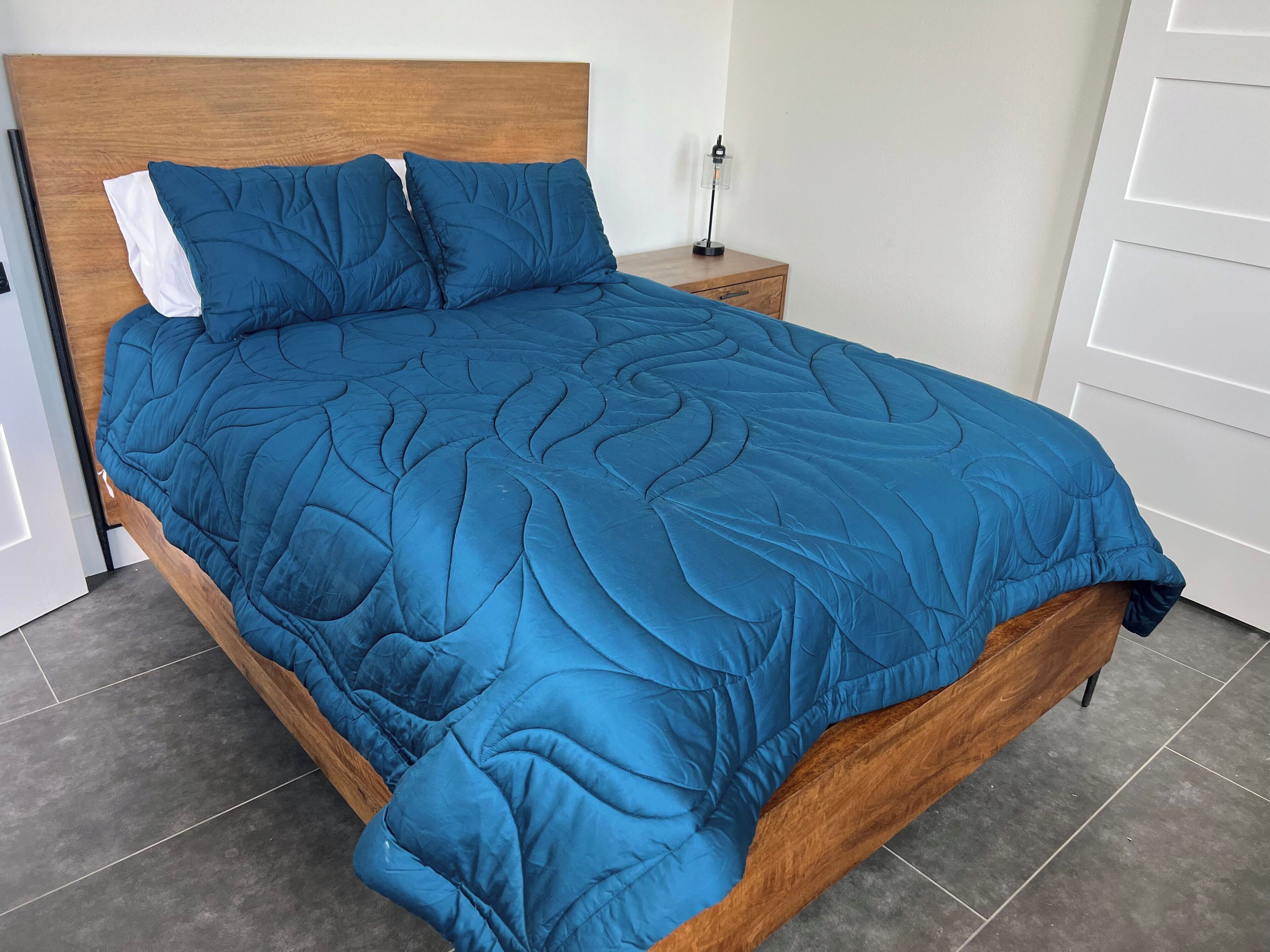 A bed with a blue quilt, a wooden headboard, and a nightstand with a lamp.