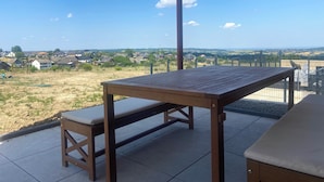 Mesa e banco de madeira numa varanda com vista para uma área residencial e colinas ao longe.