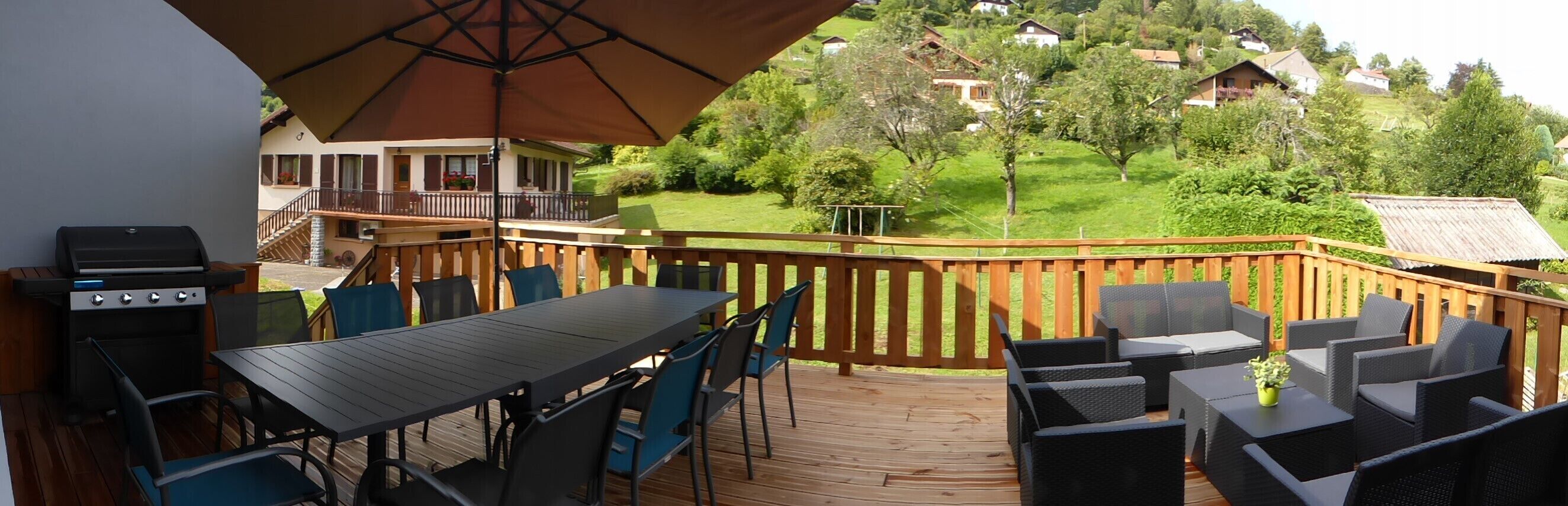 Une terrasse avec un barbecue, une table, des chaises et une vue sur un paysage verdoyant parsemé de maisons.