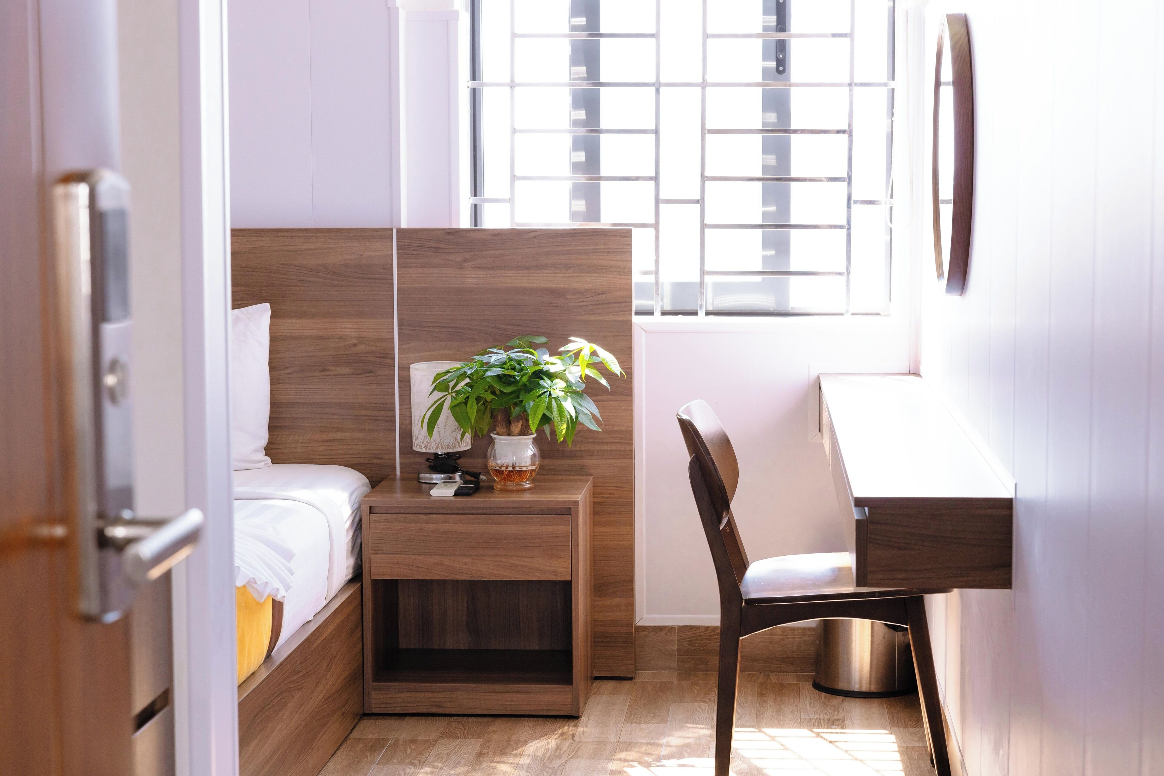 A compact bedroom with a wooden headboard, a bedside table with a plant, a desk, and a chair.