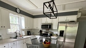 A modern kitchen with white cabinets, stainless steel appliances, a dining table with chairs, and a fruit bowl on the table. - Cozy cabin with riverfront location, fire pit and peaceful surroundings (Ellijay)