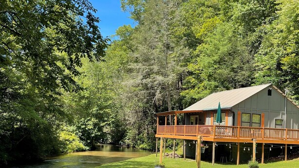 A house with a wooden deck and a covered porch, surrounded by lush greenery and a river. - Cozy cabin with riverfront location, fire pit and peaceful surroundings (Ellijay)