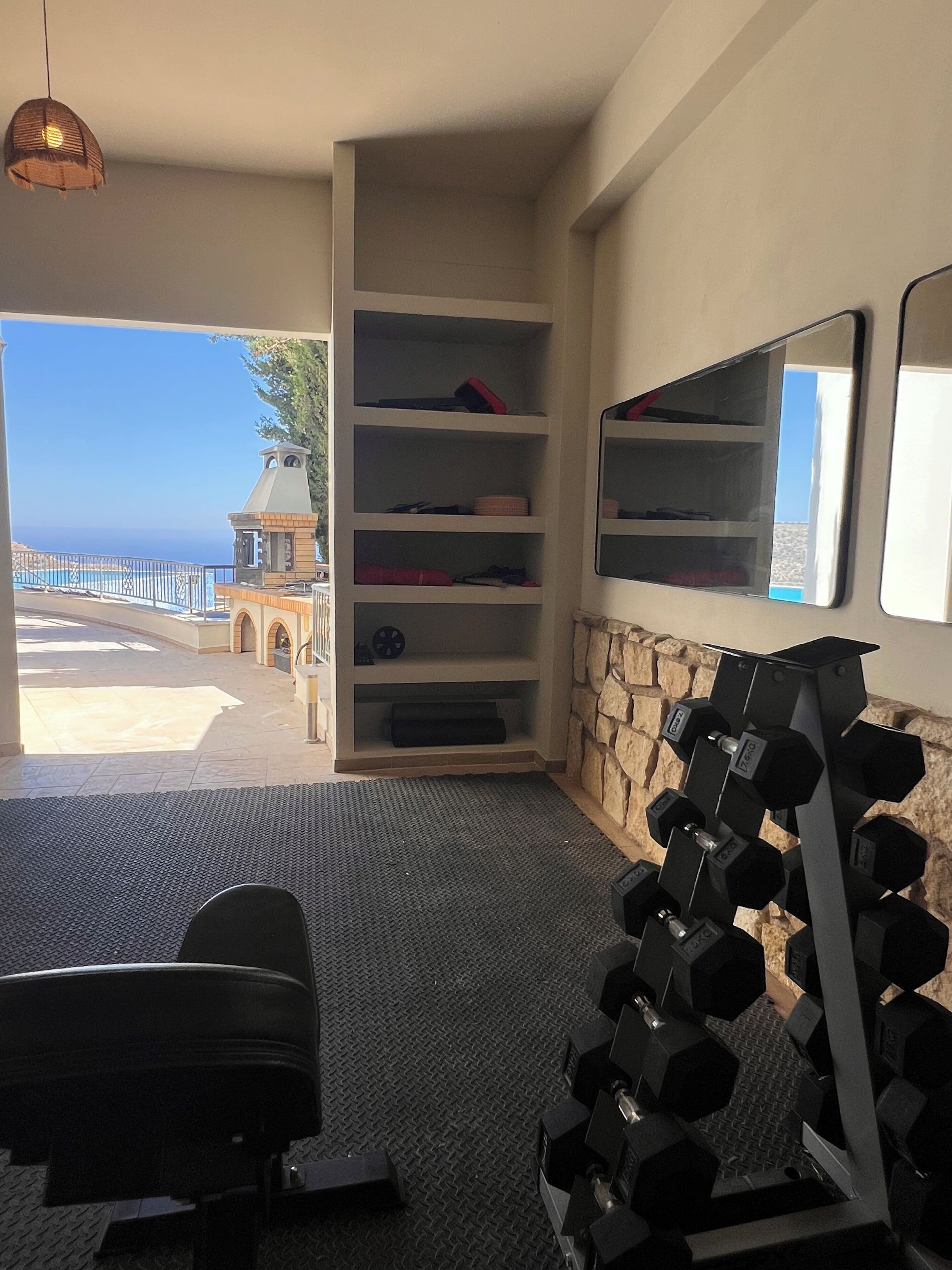 A room with a view of the sea, a stone wall, a mirror, and a set of dumbbells.