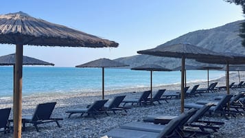Une zone aménagée au bord de la plage, avec des chaises longues et des parasols en chaume, une plage de galets et une vue dégagée sur la mer.