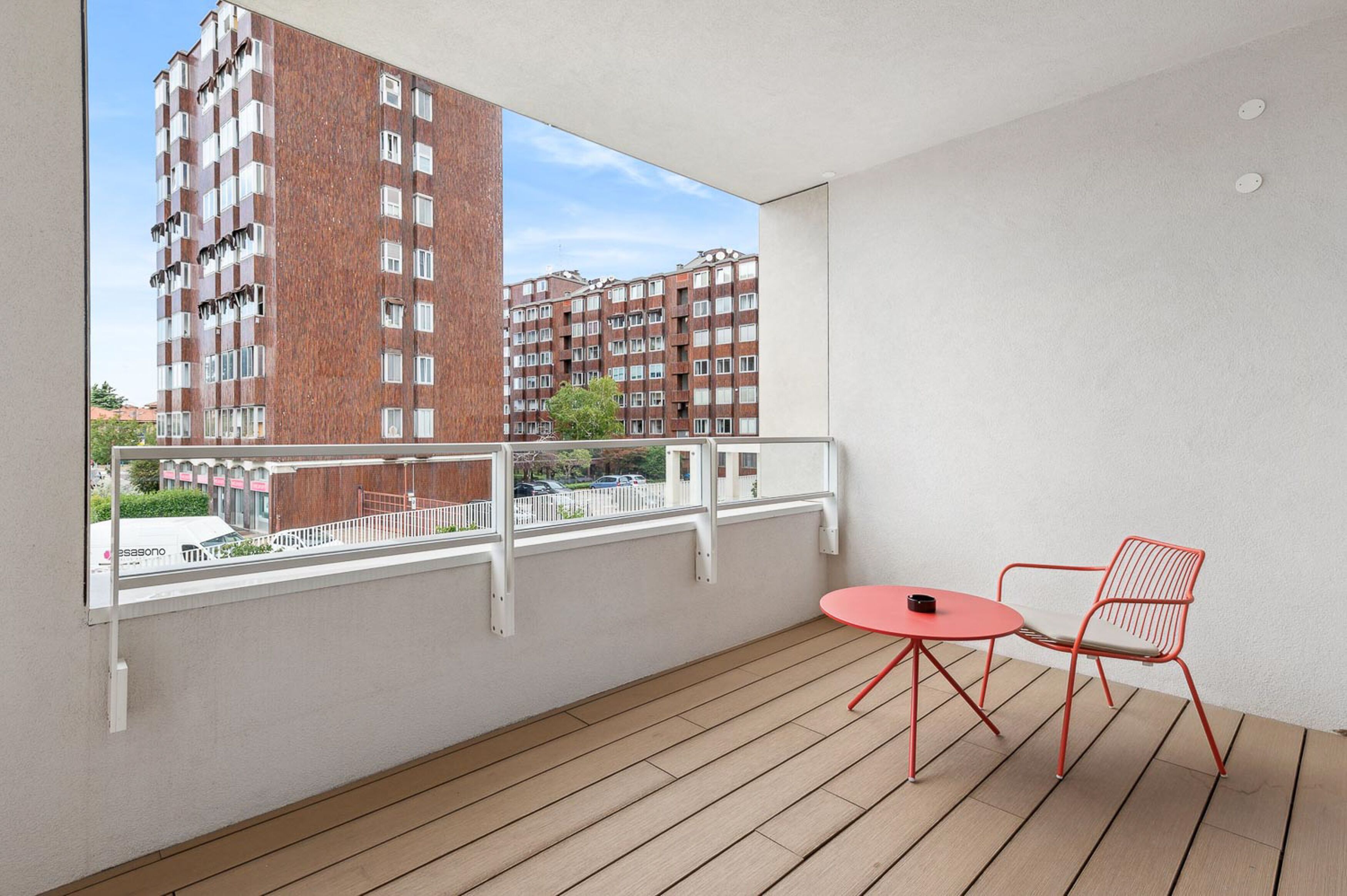 Un balcón con suelo de madera, una mesa y una silla rojas. Se observa una barandilla blanca y una vista de un edificio de ladrillo.