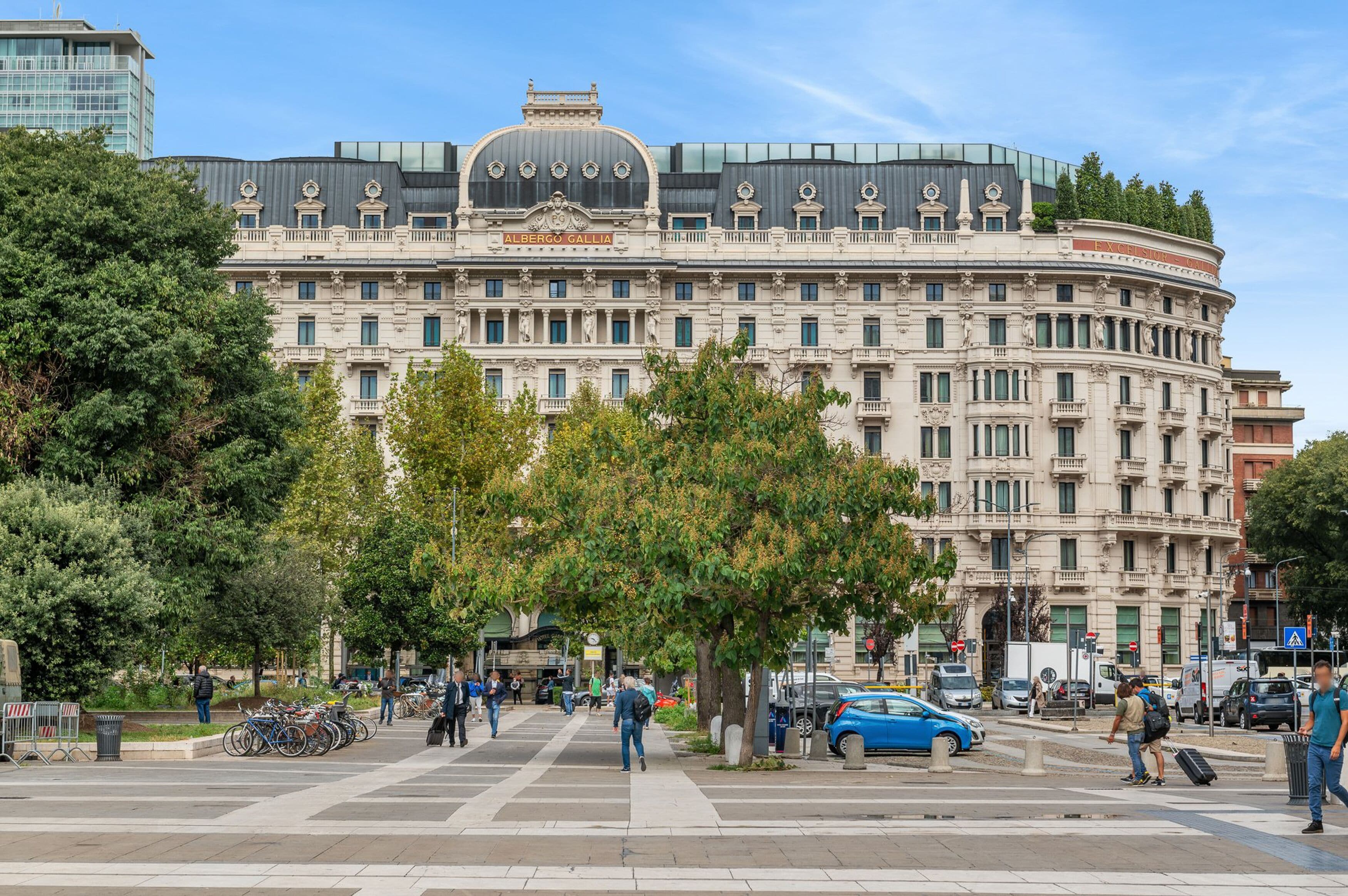 Un edificio grande e histórico con una cúpula y varios balcones, rodeado de árboles y una zona peatonal.