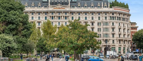 Un edificio grande e histórico con una cúpula y varios balcones, rodeado de árboles y una zona peatonal.