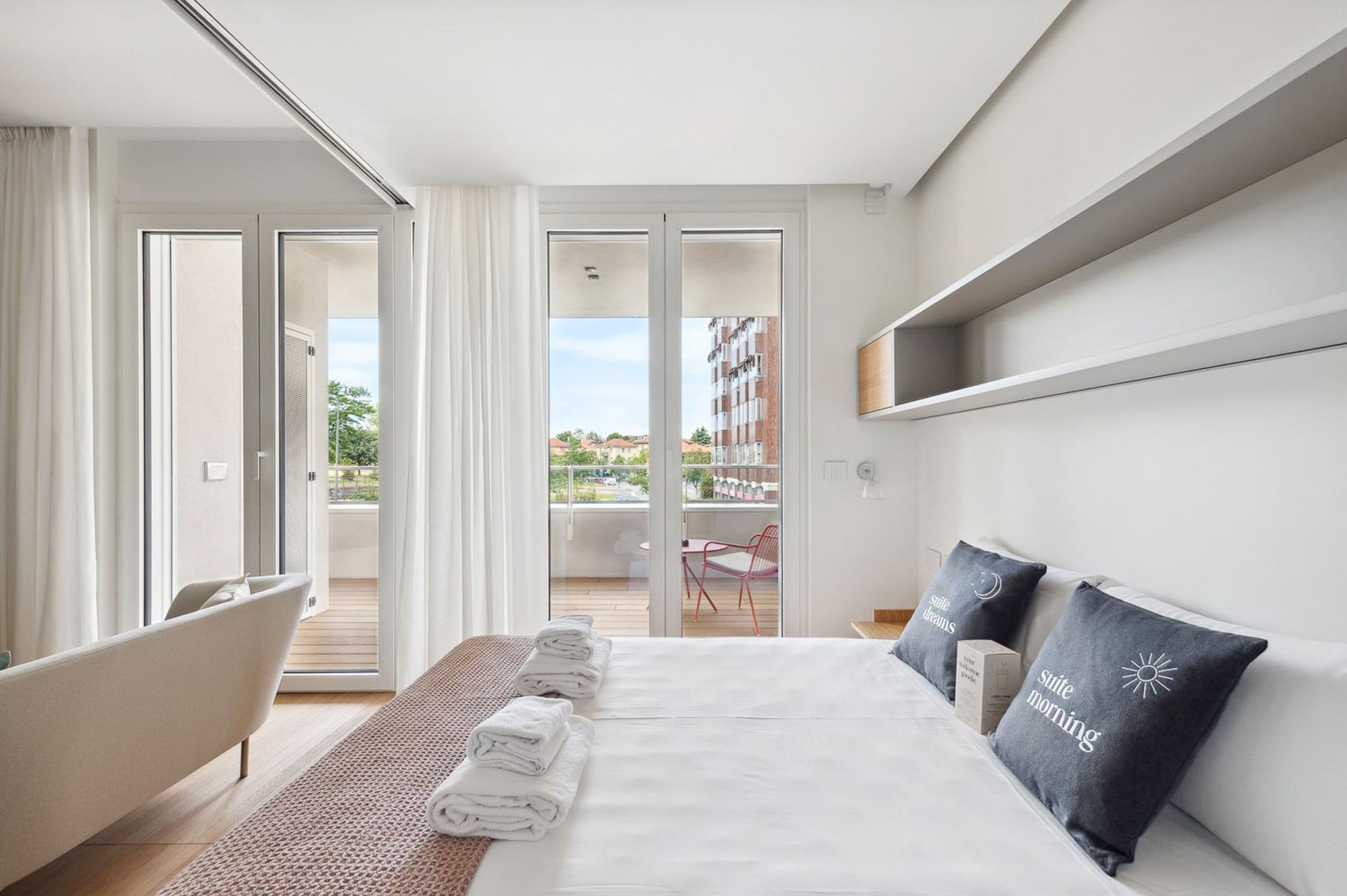 A modern hotel room with a large bed, a sitting area with a chair, a balcony with a table, and a view of the cityscape.