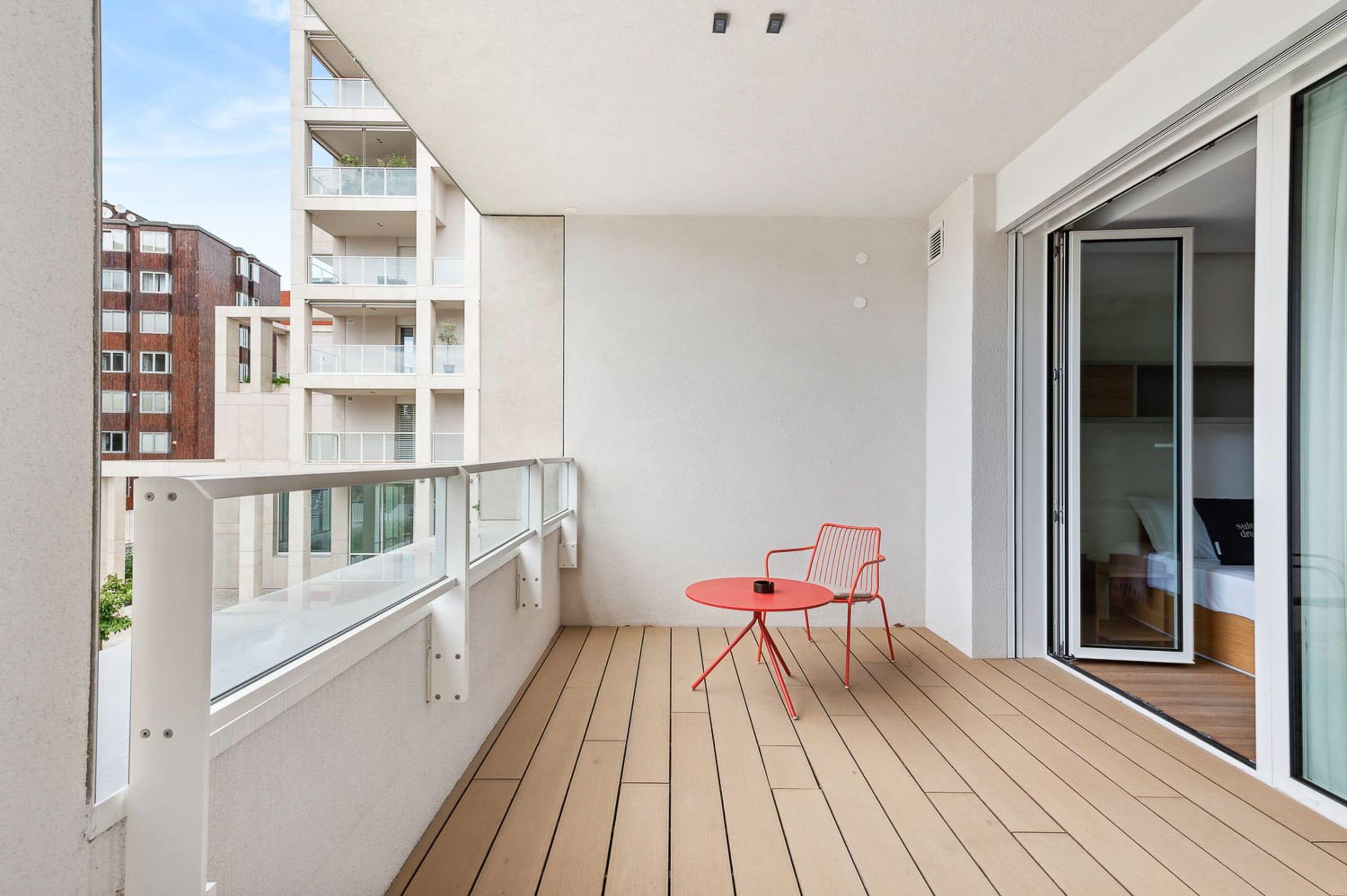 A balcony with a wooden floor, a glass door leading to a room, and a red chair.