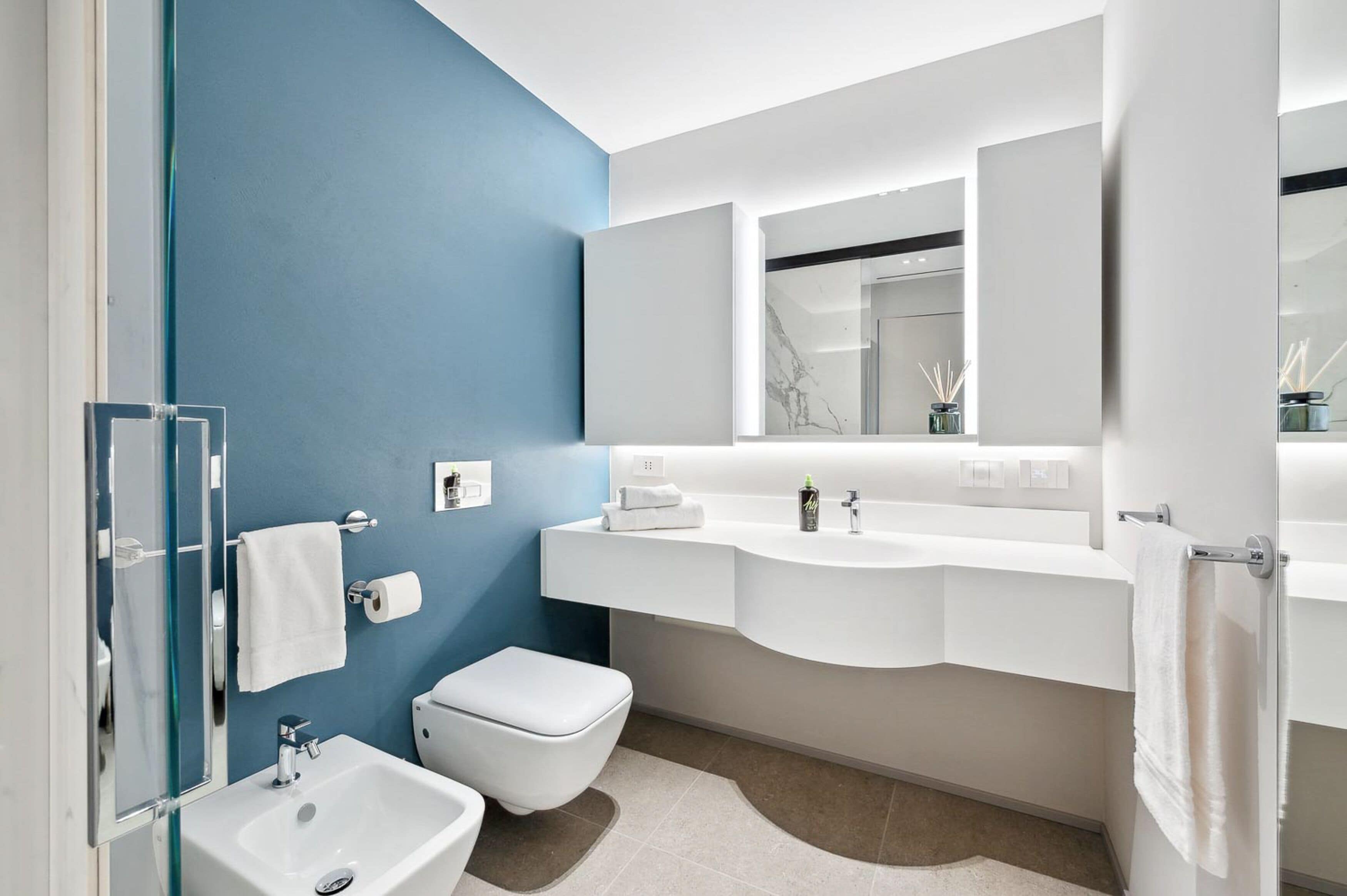 A modern bathroom with a blue accent wall, a white sink, a toilet, and a glass shower enclosure.