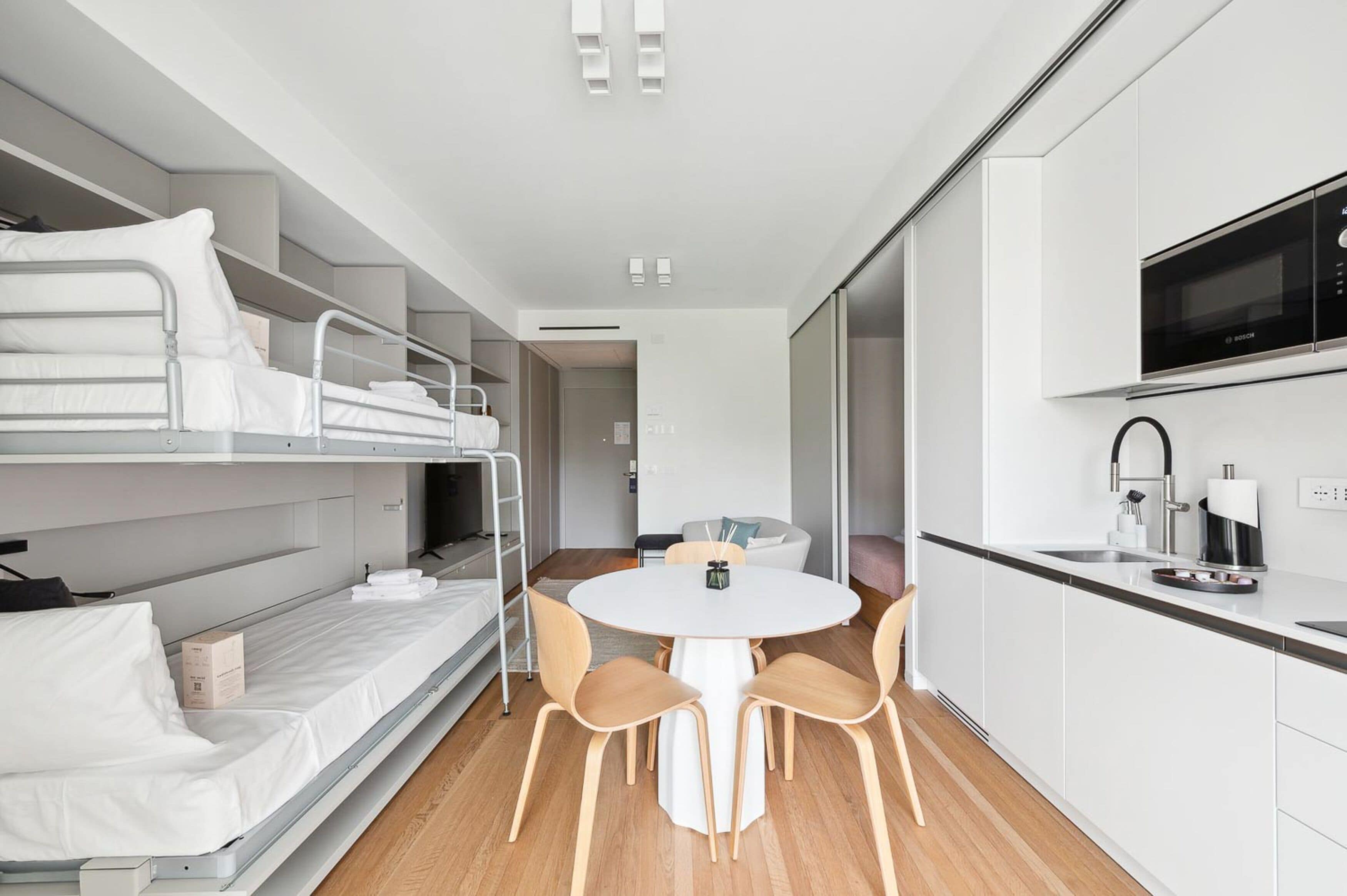 Une pièce compacte et moderne, dotée de lits superposés, d’une table de salle à manger ronde avec des chaises, d’une kitchenette équipée d’un four à micro-ondes et d’un petit coin salon.