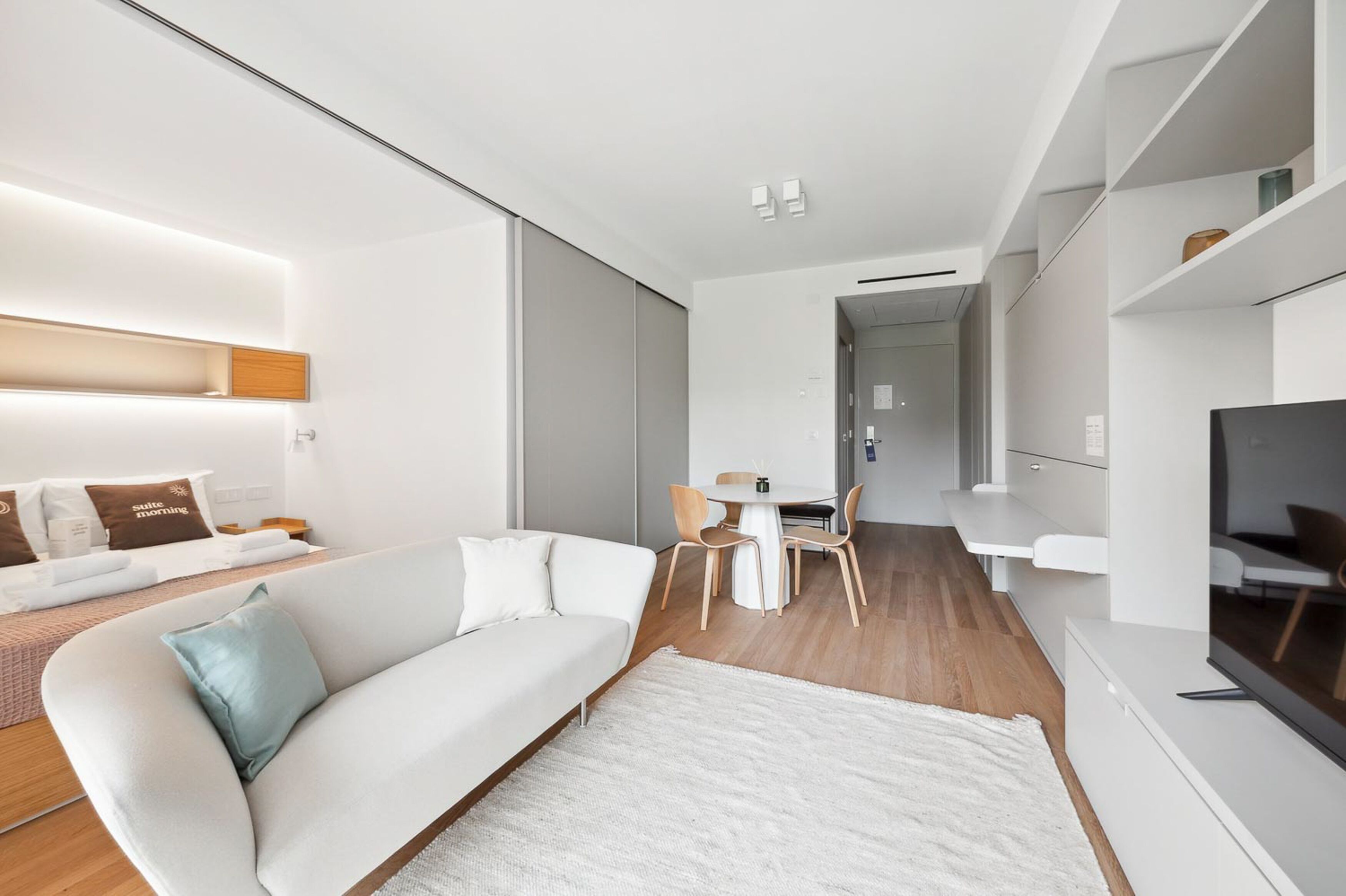 Una sala de estar moderna con un sofá blanco, una mesa de comedor con sillas, un televisor de pantalla plana y un ventanal grande.