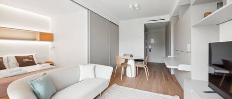 A modern living room with a white sofa, a dining table with chairs, a flat-screen TV, and a large window.