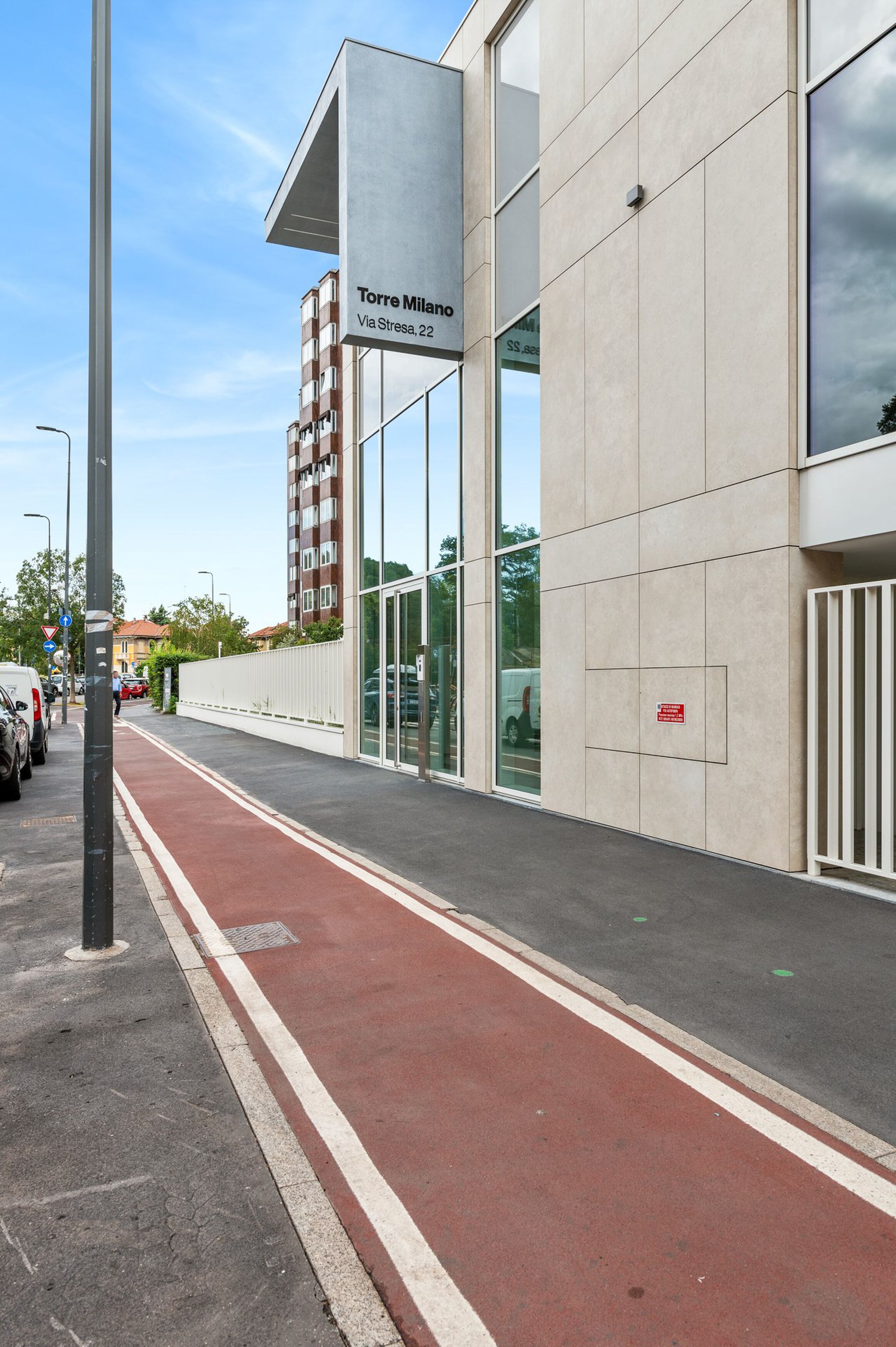 A modern building with a red bike lane, a sign reading "Torre Milano Via Stresa, 22", and a white fence.