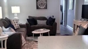 En moderne stue med sofa, sofabord og en lille skammel. Der er ventilator i loftet, en lampe og et fjernsyn på et stativ.