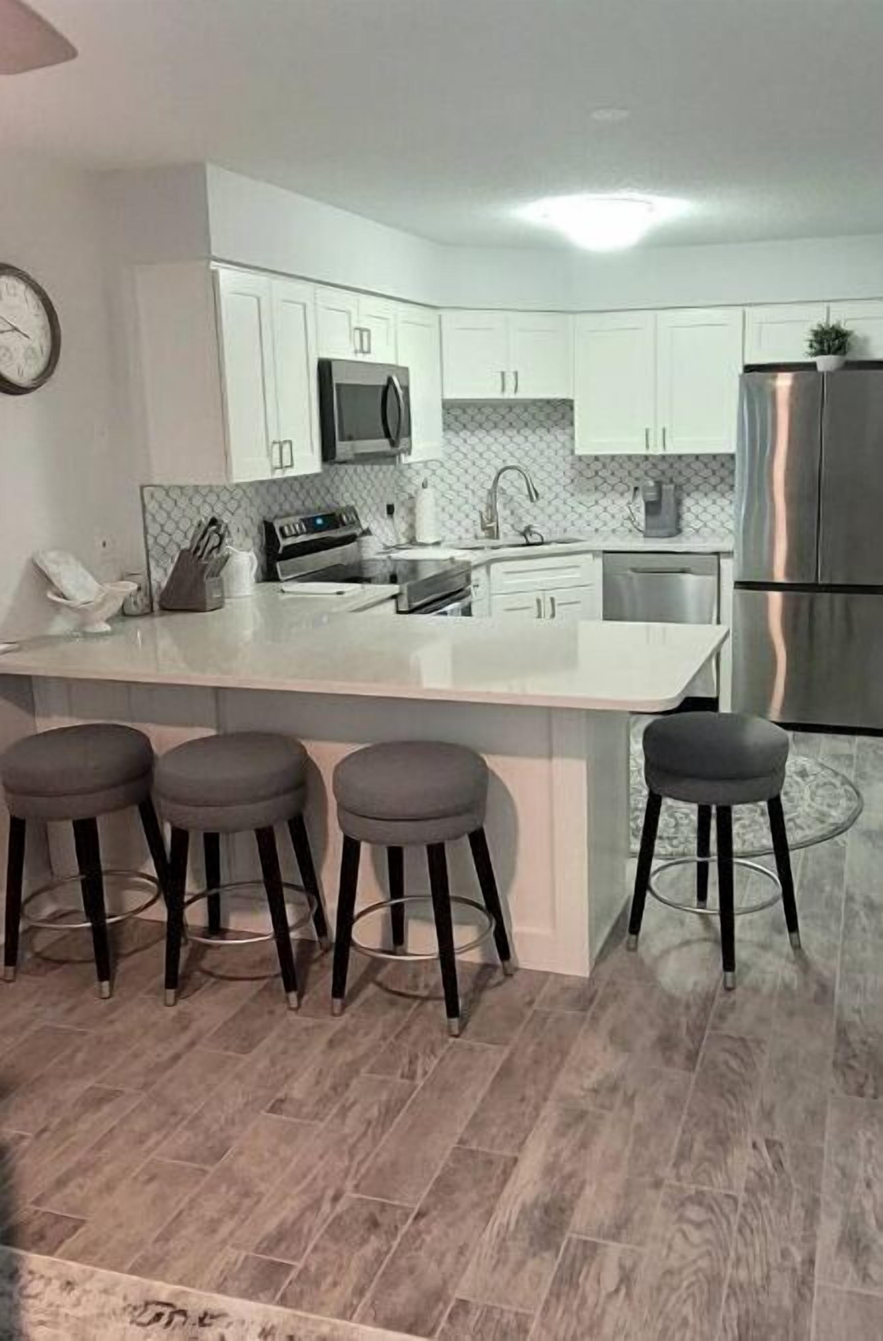 A modern kitchen with white cabinets, a stainless steel refrigerator, a microwave, and a sink. There are three bar stools and a clock on the wall.