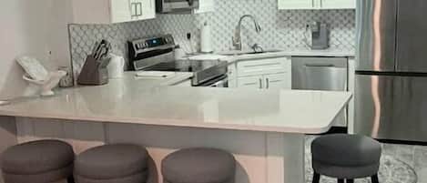 A modern kitchen with white cabinets, a stainless steel refrigerator, a microwave, and a sink. There are three bar stools and a clock on the wall.