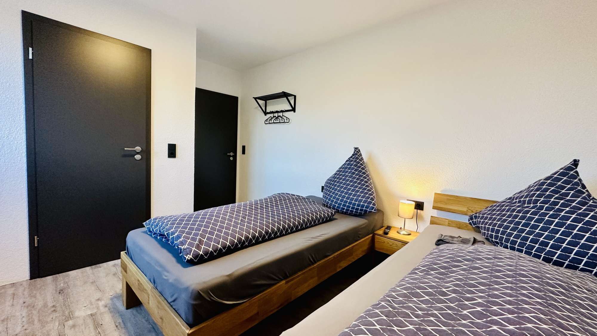 Une petite pièce propre, avec deux lits, un plancher en bois, une porte noire et un luminaire fixé au mur.