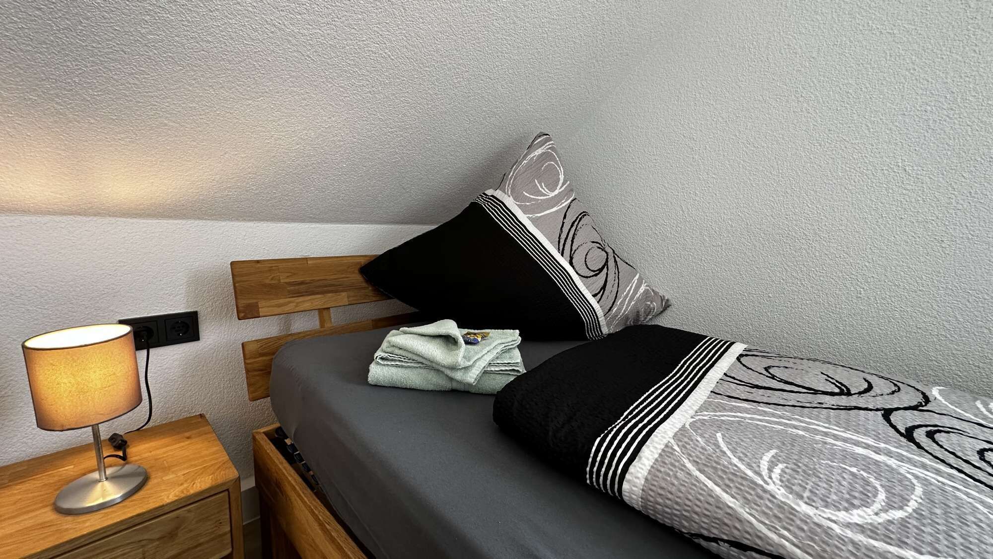 Una cama bien hecha con una colcha gris y negra estampada, una toalla doblada y una almohada negra con un diseño blanco. Hay una mesita de noche de madera con una lámpara y un enchufe.