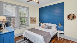 A bedroom with a bed, a ceiling fan, a window with blinds, a wall-mounted lamp, and a small table.