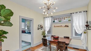 A dining area with a wooden table, chairs, a chandelier, and a view of the kitchen through a window.