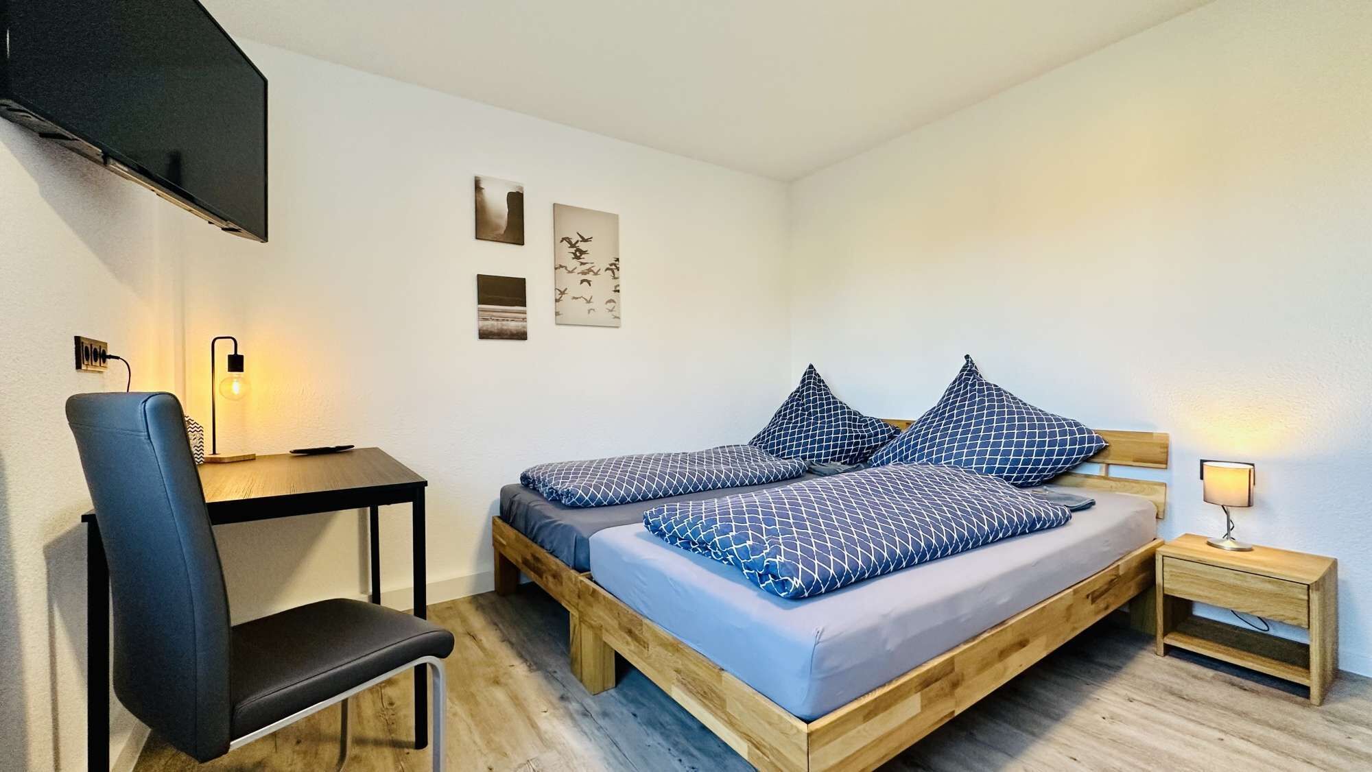 A bedroom with a wooden bed, two pillows, a desk, a chair, and a TV mounted on the wall.