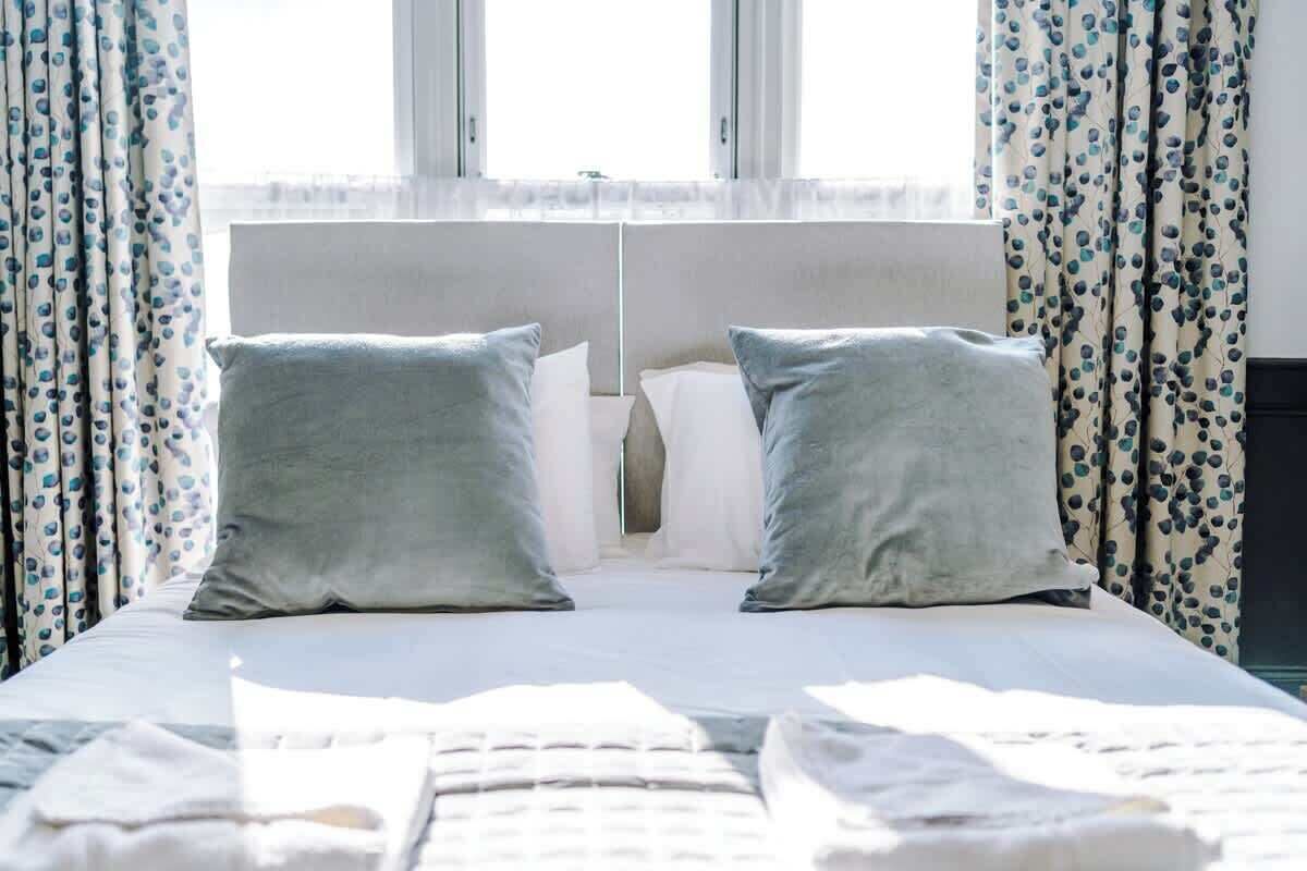 A neatly made bed with two pillows, a window with curtains, and a headboard.