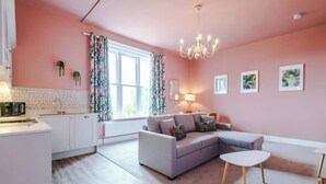 Et rosa-tema stue med sofa, to sofaborde, en lysekrone og et vindue med gardiner.