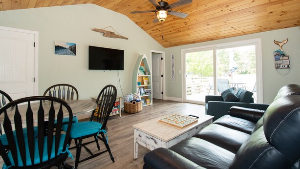A living room with a wooden ceiling, a ceiling fan, a TV mounted on the wall, a dining table with chairs, a coffee table, and a sofa.