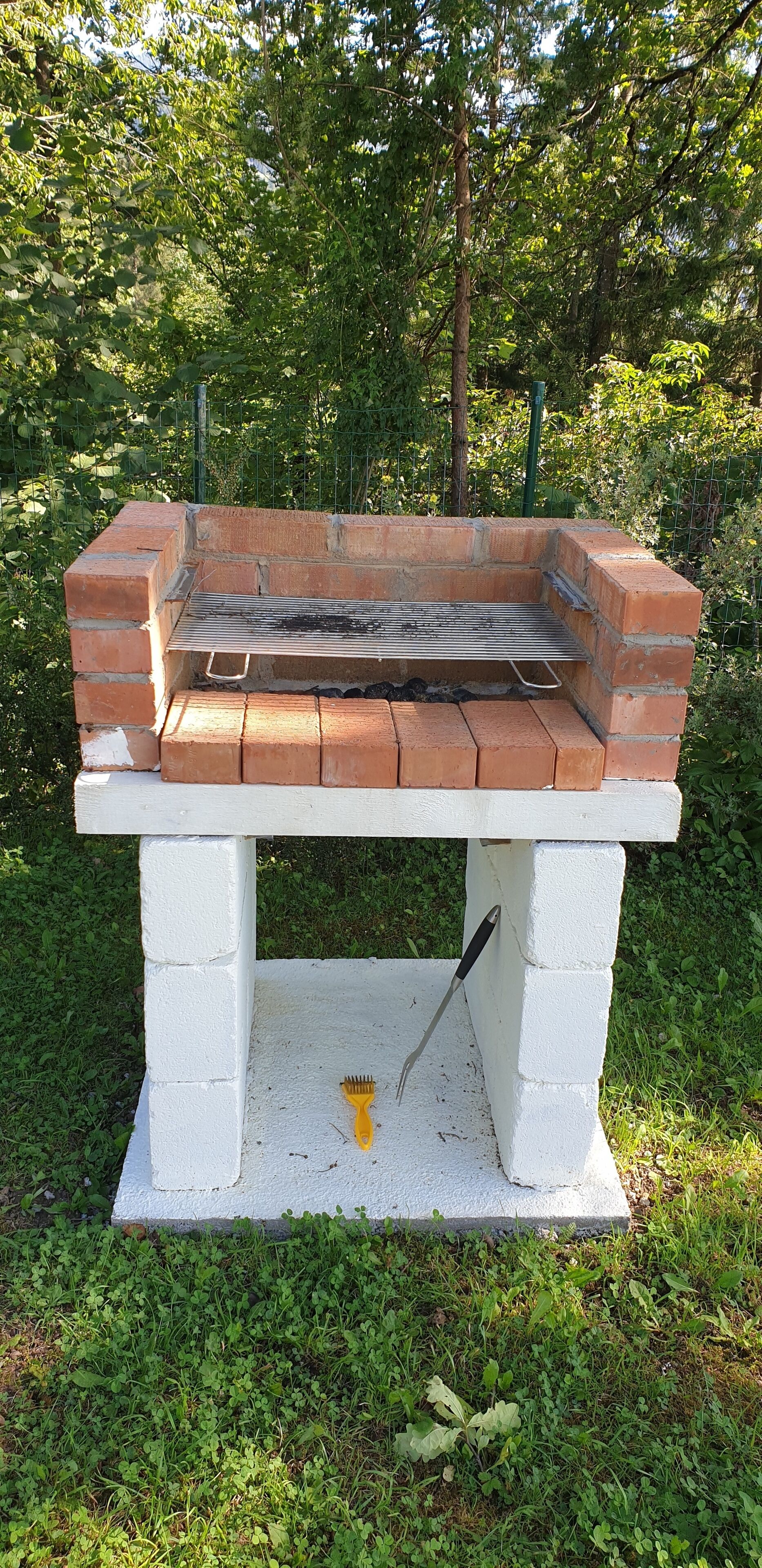 A brick barbecue grill with a metal grate, set on a white concrete base, surrounded by greenery.