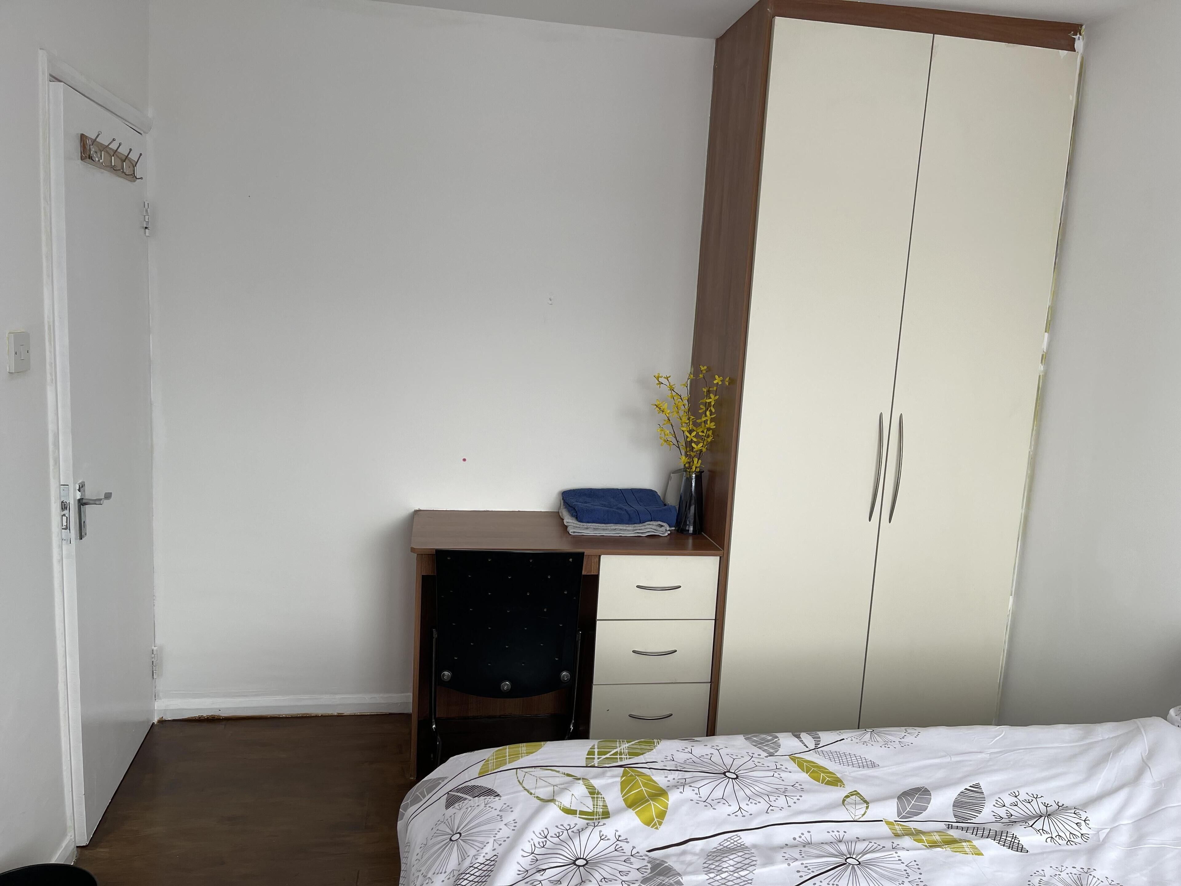 A bedroom with a bed, a desk, a wardrobe, and a small table with a vase of flowers.