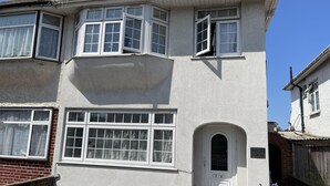 Una casa de dos pisos con un ventanal saliente, una puerta principal con felpudo y un muro de ladrillo.