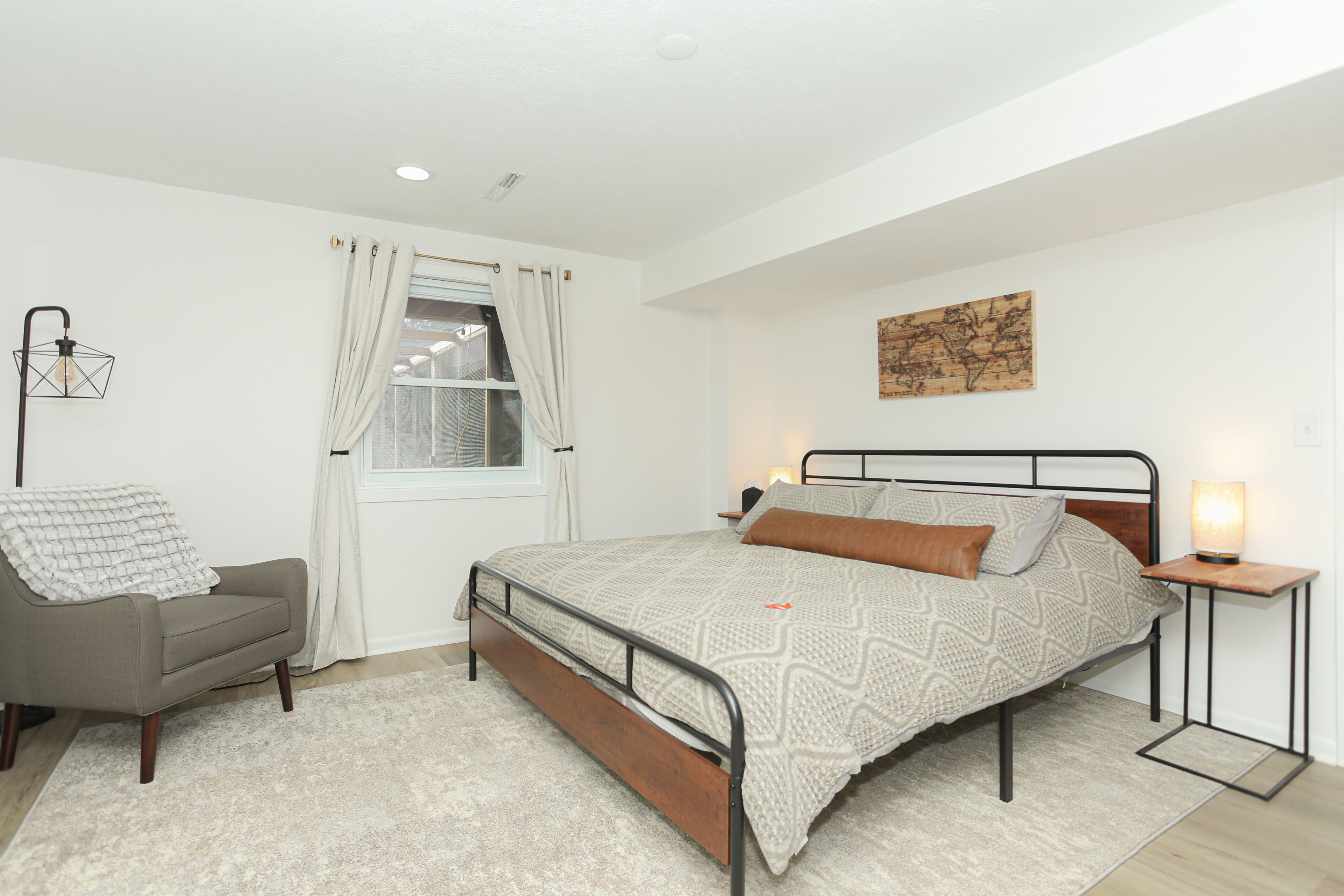 Un dormitorio con cama, una silla, una mesita de noche, una lámpara y una ventana con cortinas.