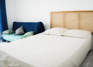 Een bed met witte beddengoed, een blauwe bank met opvallende kussens, en een geweven hoofdbord.